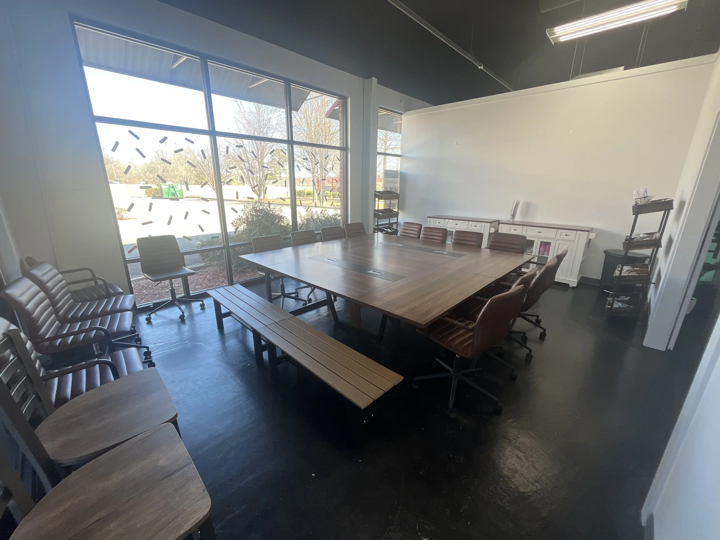 A conference room with a large wooden table, surrounded by brown padded chairs with armrests. Large windows on one side allow natural light, and outside there are trees and a parking lot. White cabinets and black shelves are along the walls, and the ceiling has black panels and fluorescent lights.