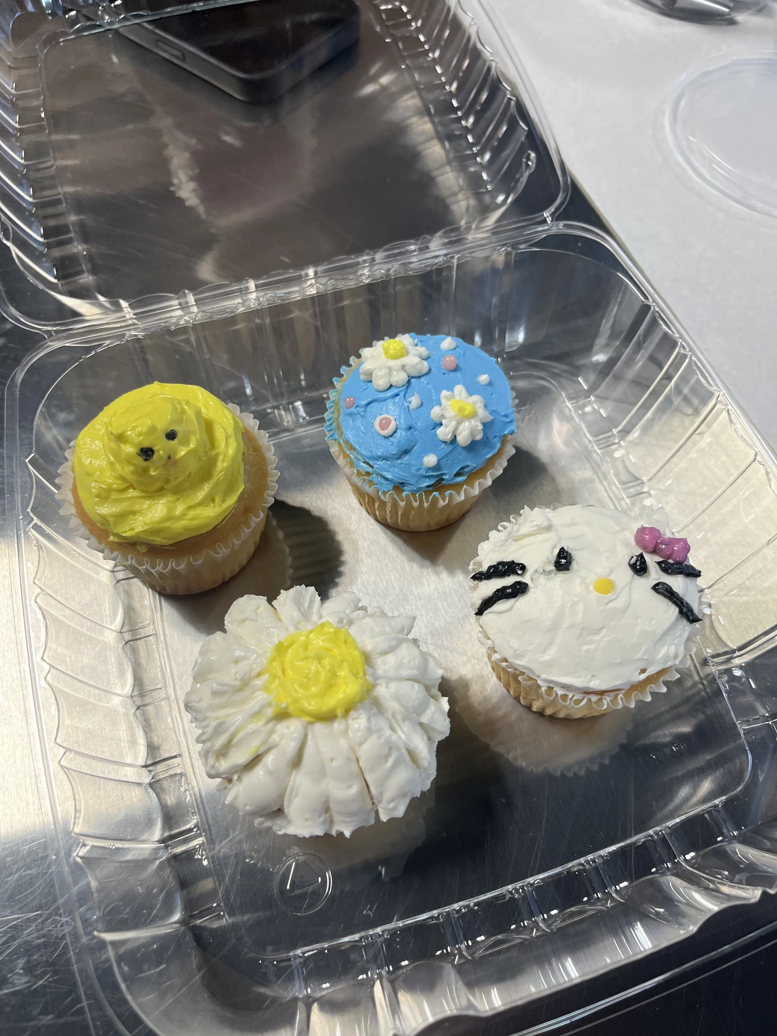 A plastic container with decorated cupcakes. The cupcakes have various designs: one with yellow frosting resembling a chick, one with blue frosting decorated with flowers and small pink and white balls, and another with white frosting decorated with black and pink icing resembling a face or animal.