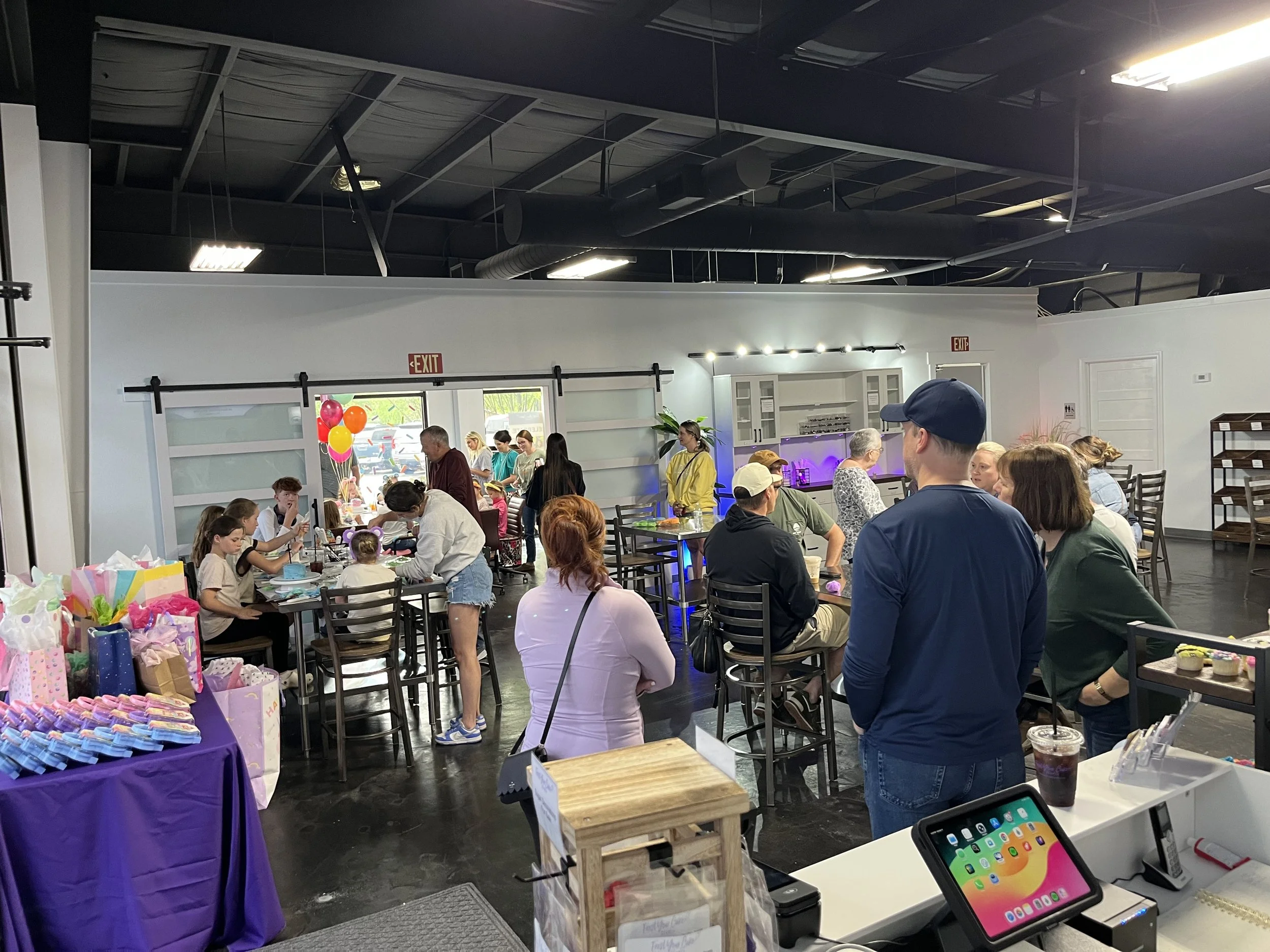 A gathering inside a modern, industrial-style space decorated for a celebration, with people sitting at tables, kids and adults, some standing, and tables with party gifts, balloons, and colorful decorations.
