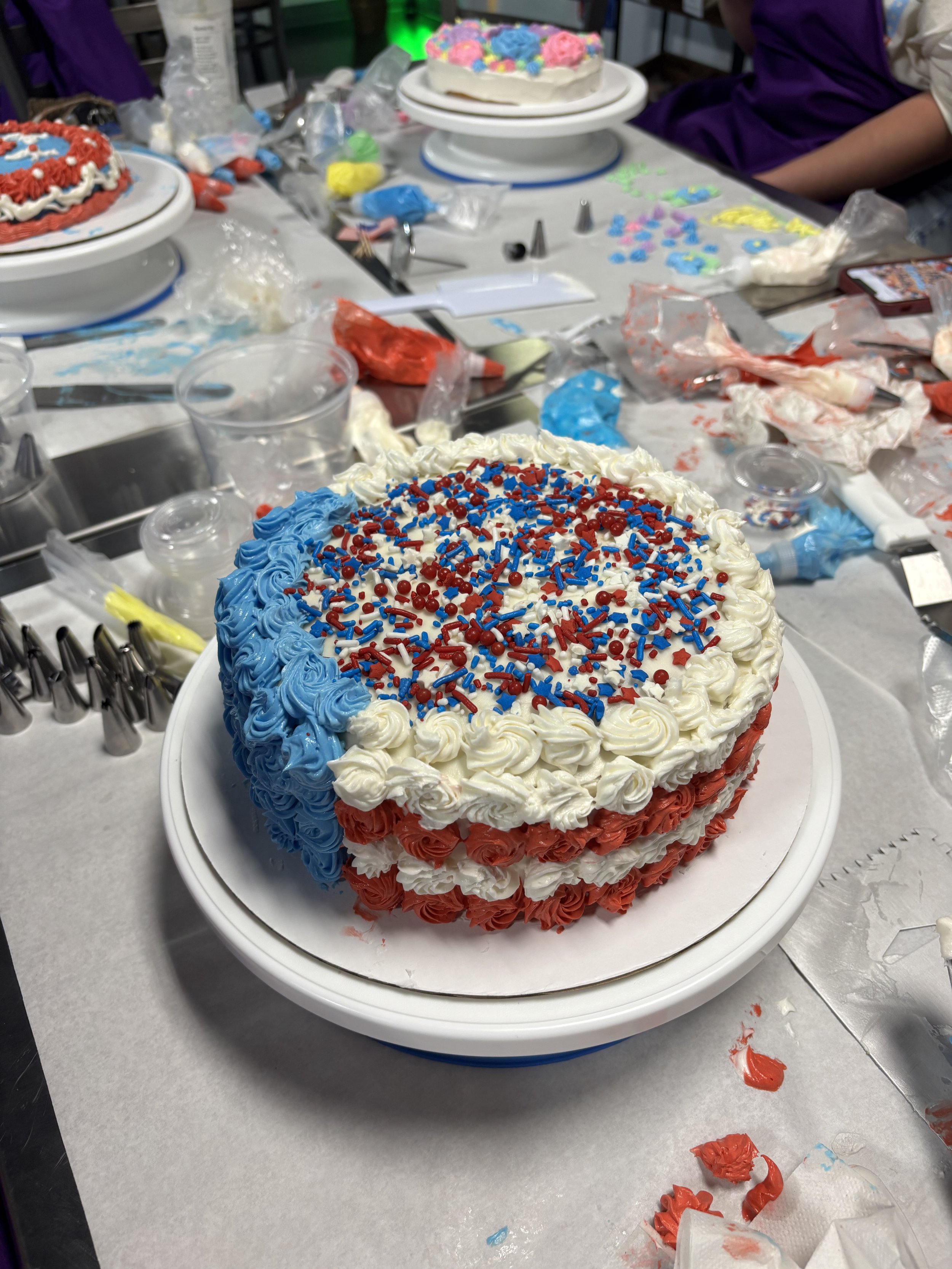 Decorated birthday cake with red, white, and blue frosting and sprinkles, sitting on a white plate on a cluttered table.