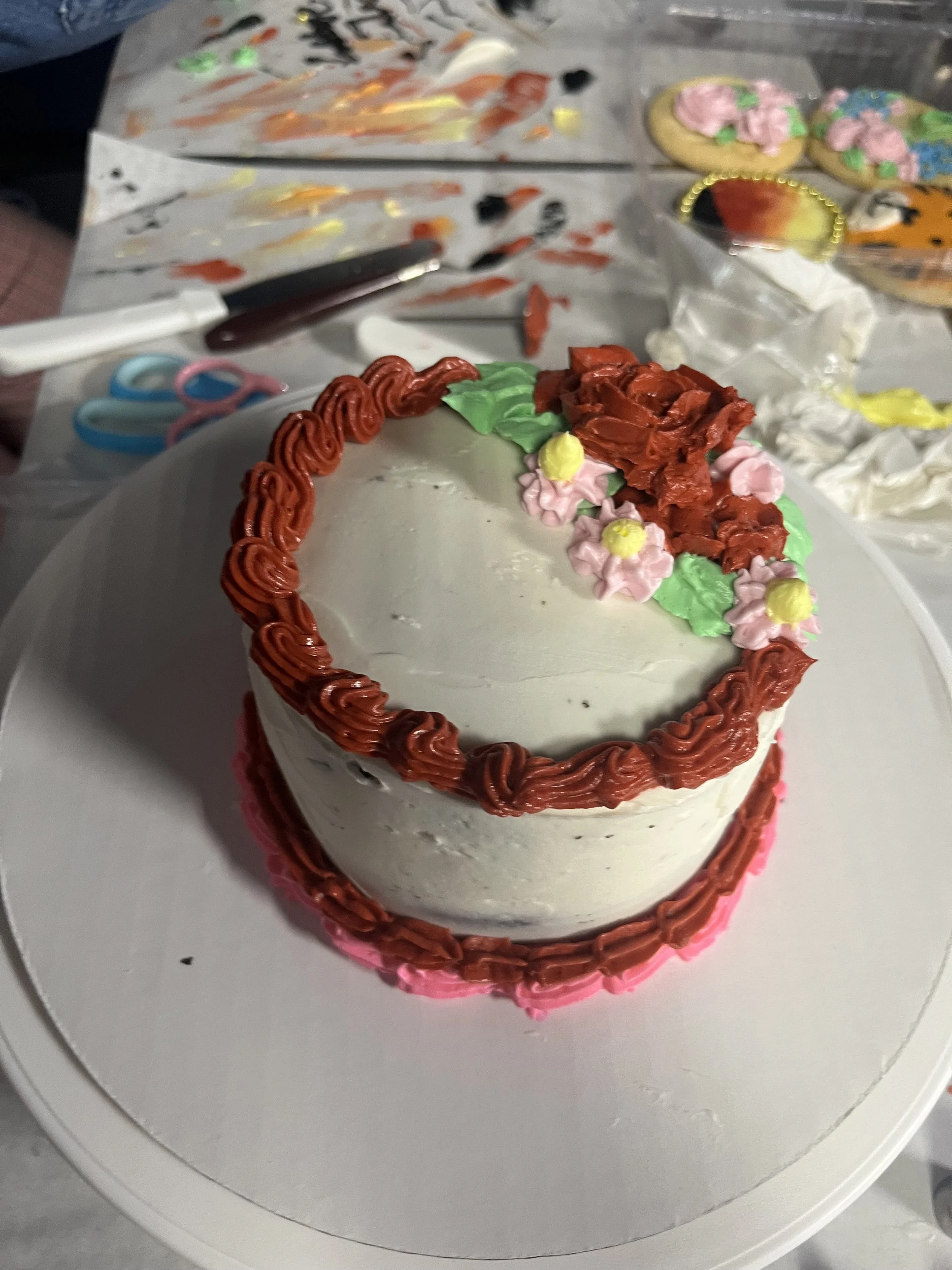 Decorative cake with white frosting, red piping around the edges, pink, green, and yellow floral designs on top.