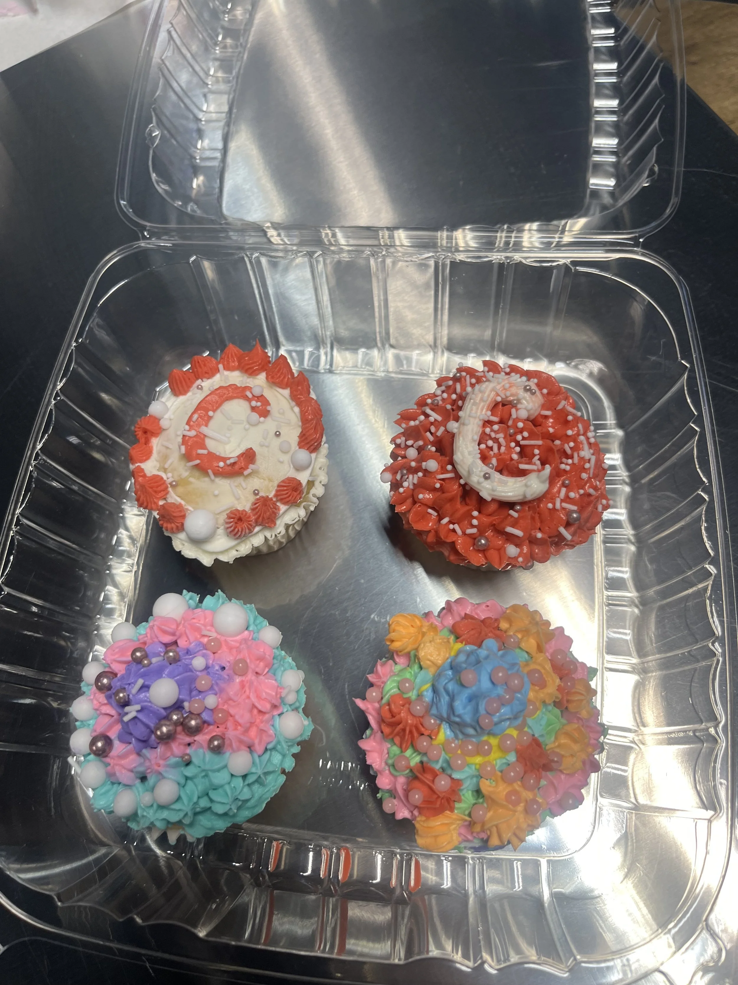 Colorful, decorated cupcakes inside a clear plastic container with a hinged lid, featuring various sprinkles and frosting decorations.