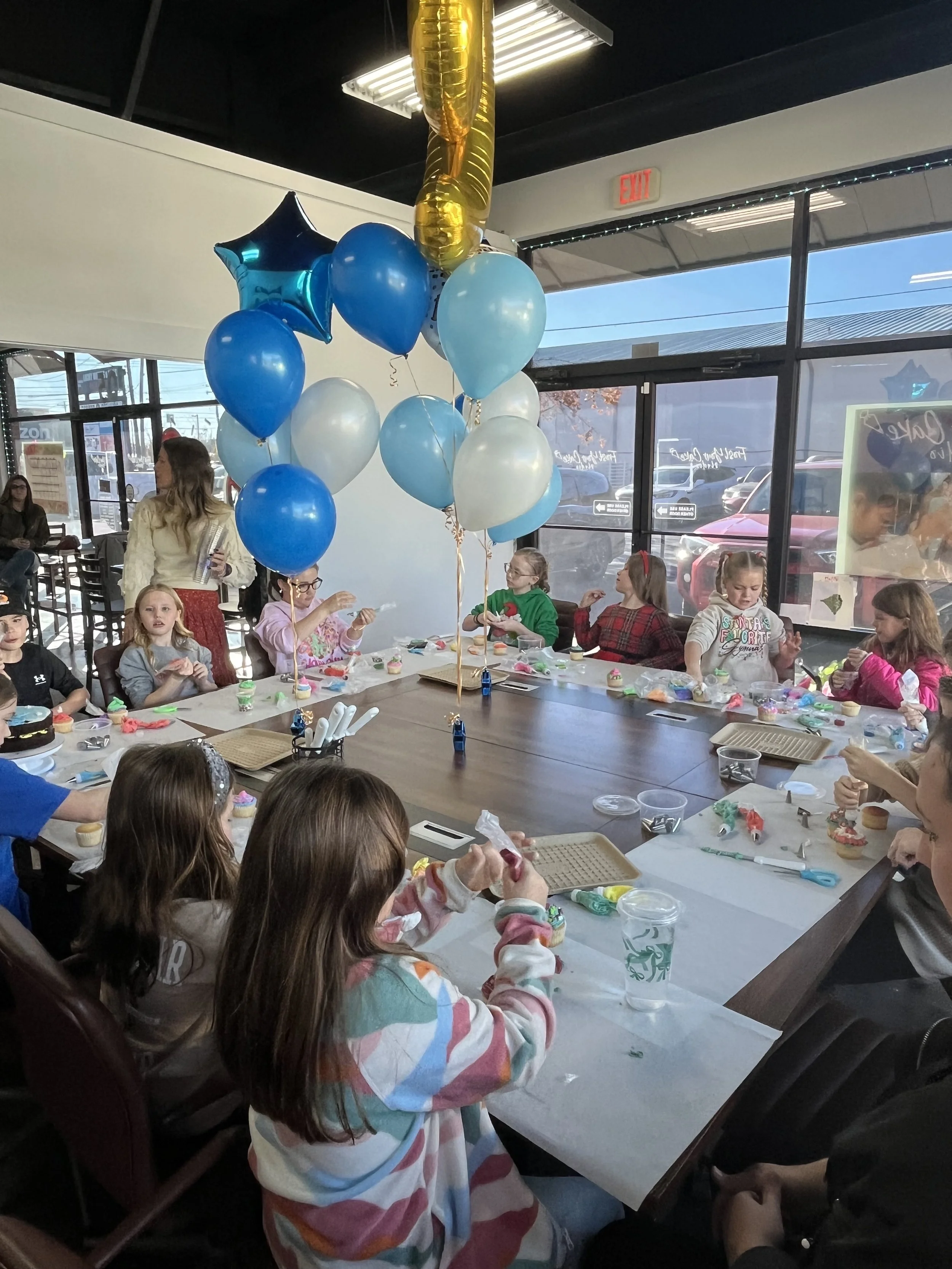 Kids and adults at a birthday party decorating cupcakes at a long table covered with a white tablecloth, balloons hanging from the ceiling, large windows in the background showing a parking lot and trees.