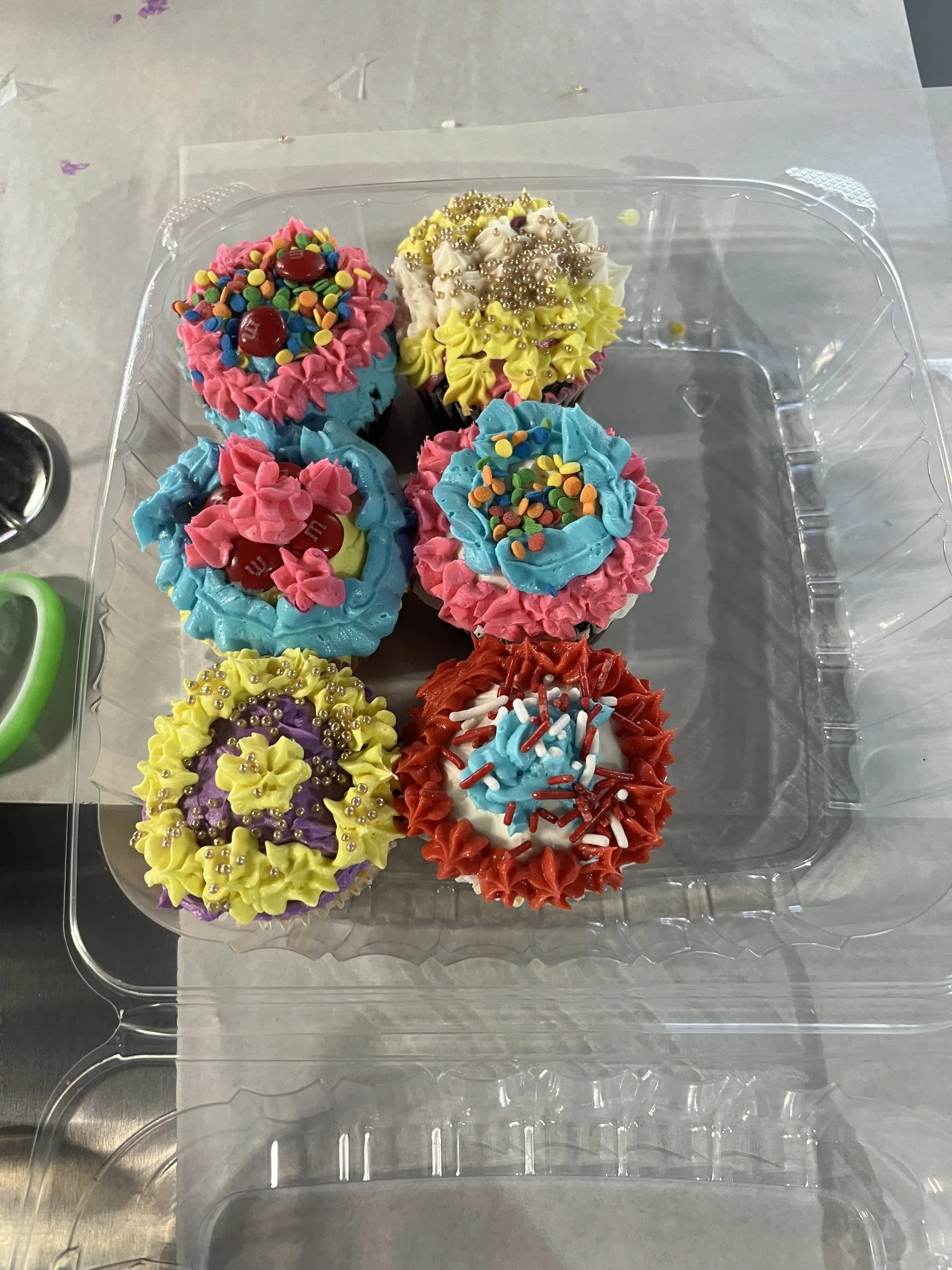 A plastic container holding five colorful cupcakes decorated with multicolored sprinkles, pearls, and icing in pink, yellow, blue, purple, and red.
