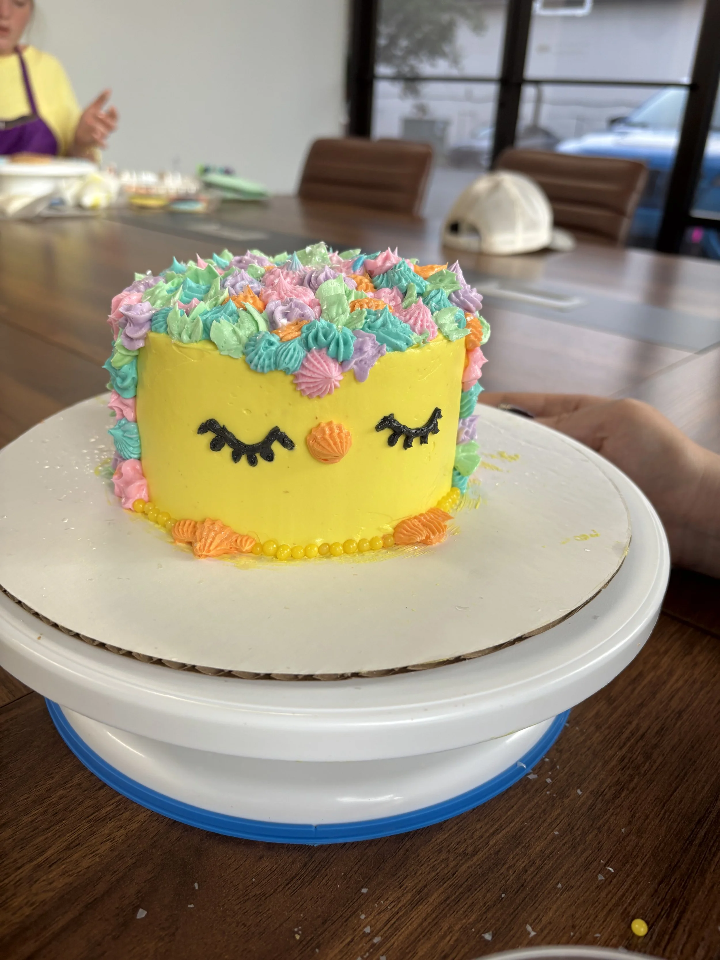 A colorful birthday cake with yellow frosting decorated with multicolored piped icing in the shape of closed eyes and a beak, resembling a bird or chick, placed on a white cake board.