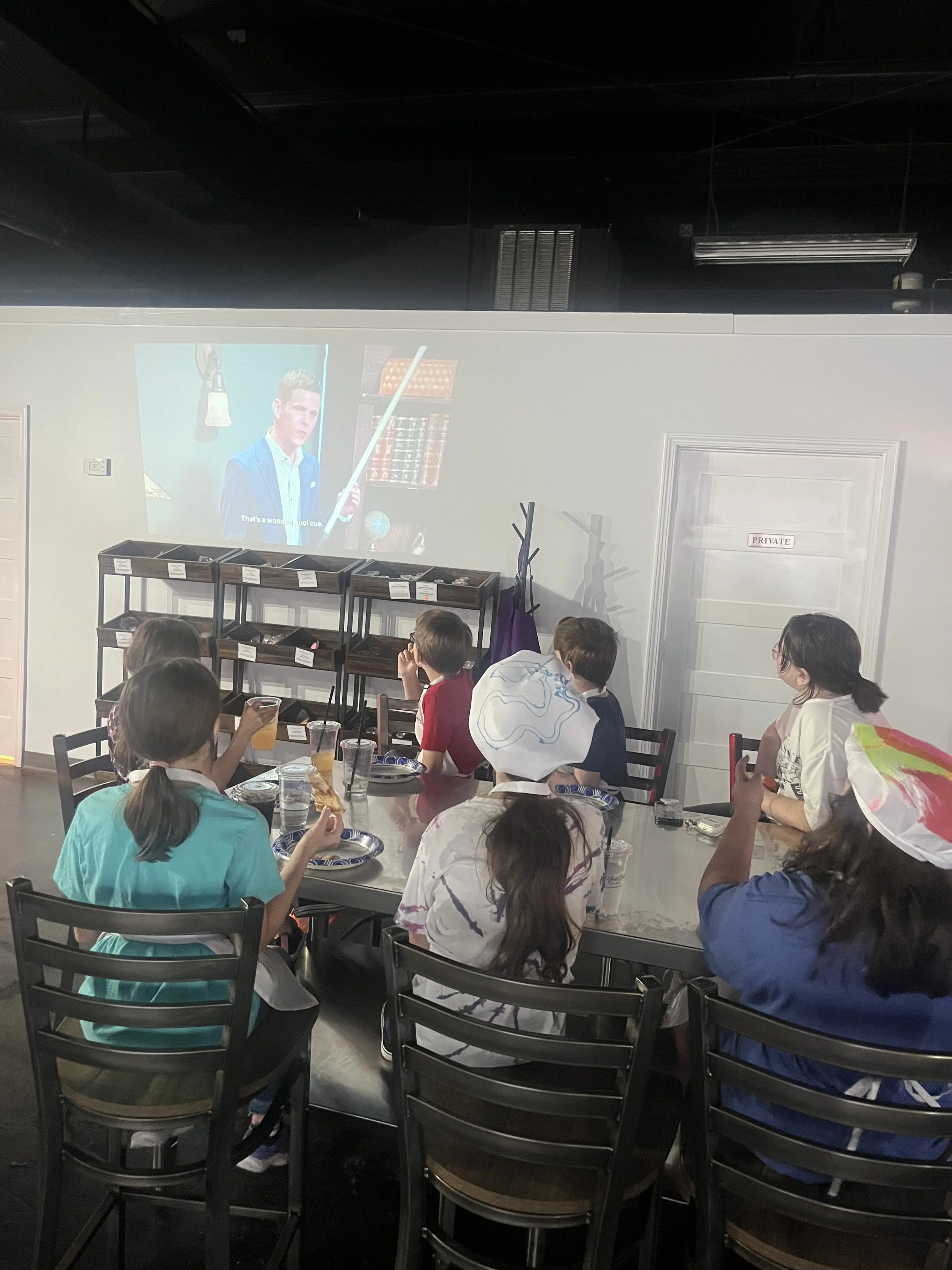 Children sitting at a dining table in a room watching a presentation on a screen, with some children drinking beverages and one wearing a chef's hat.