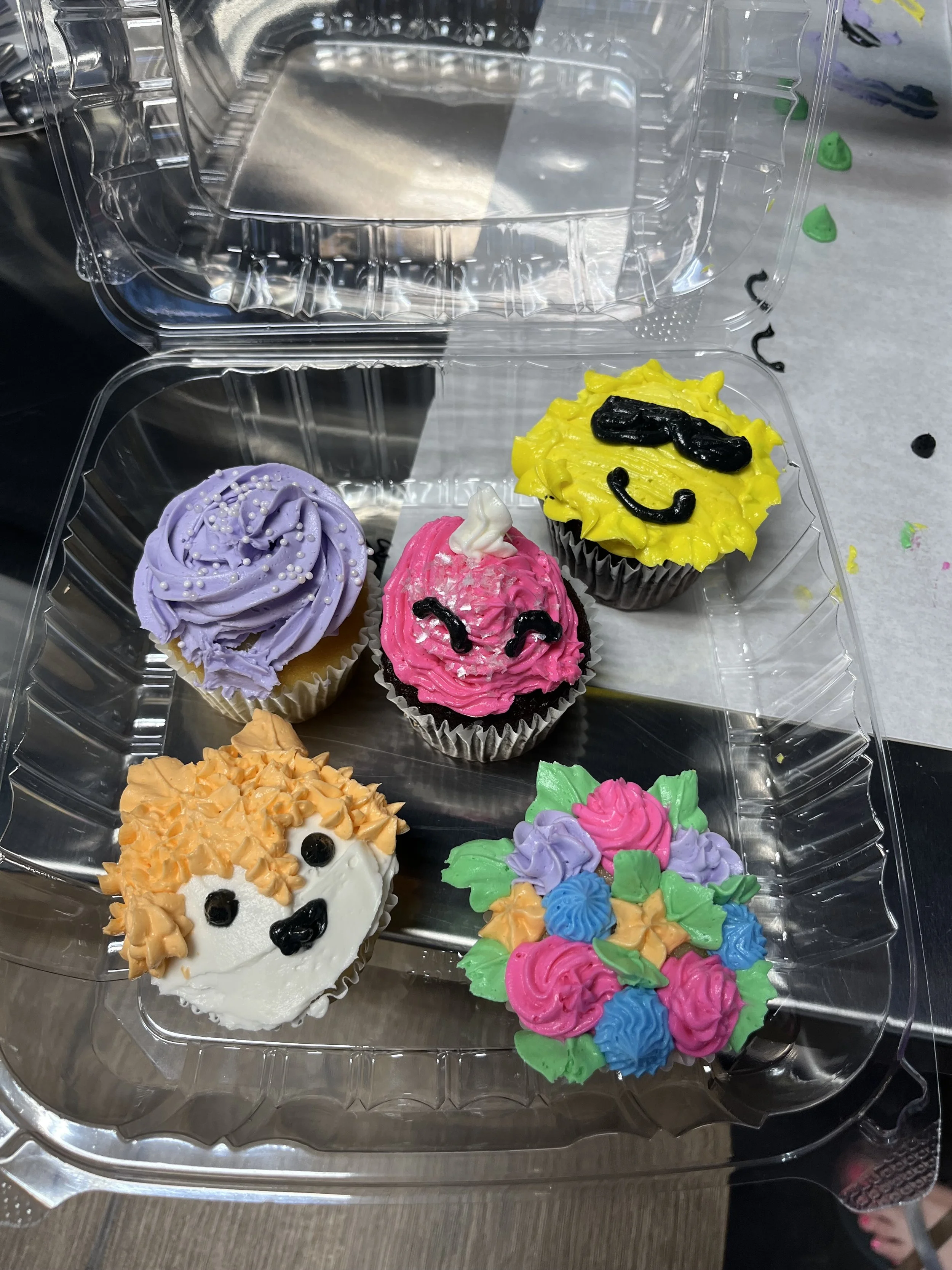 Colorful cupcakes with various decorations including a yellow smiling face, a unicorn, and floral designs in a plastic container.