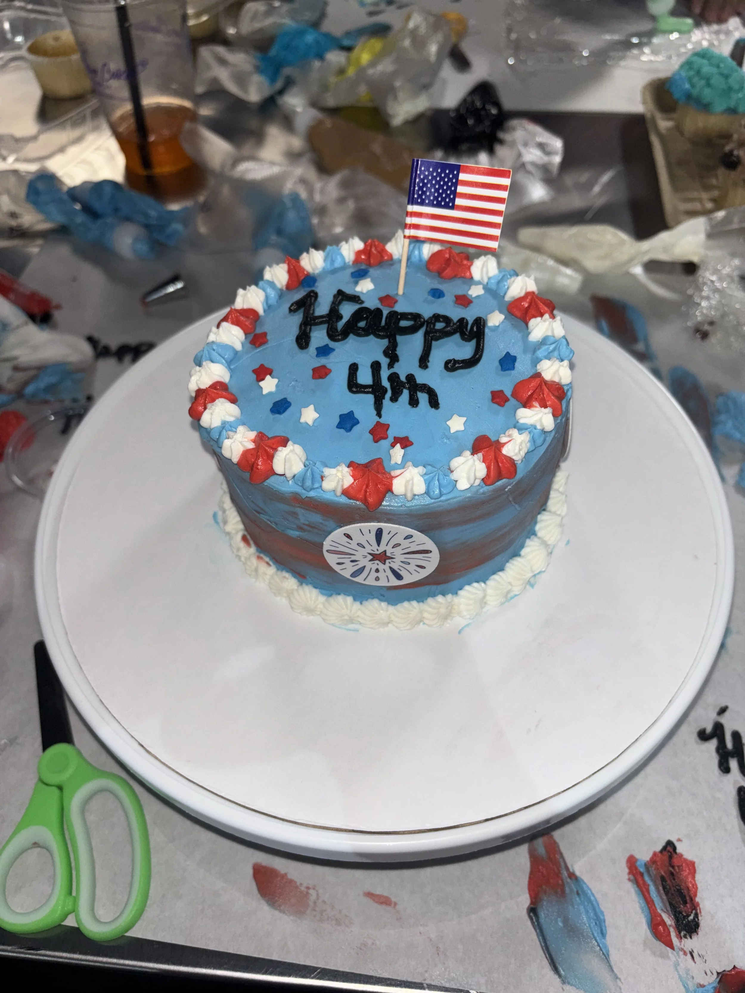 A birthday cake with blue icing, red, white, and blue star-shaped sprinkles, and an American flag topper. The cake has the words 'Happy 4th' written on it in black icing. The cake is on a white cake stand, and there are various decorating tools and supplies scattered around on the table surface.