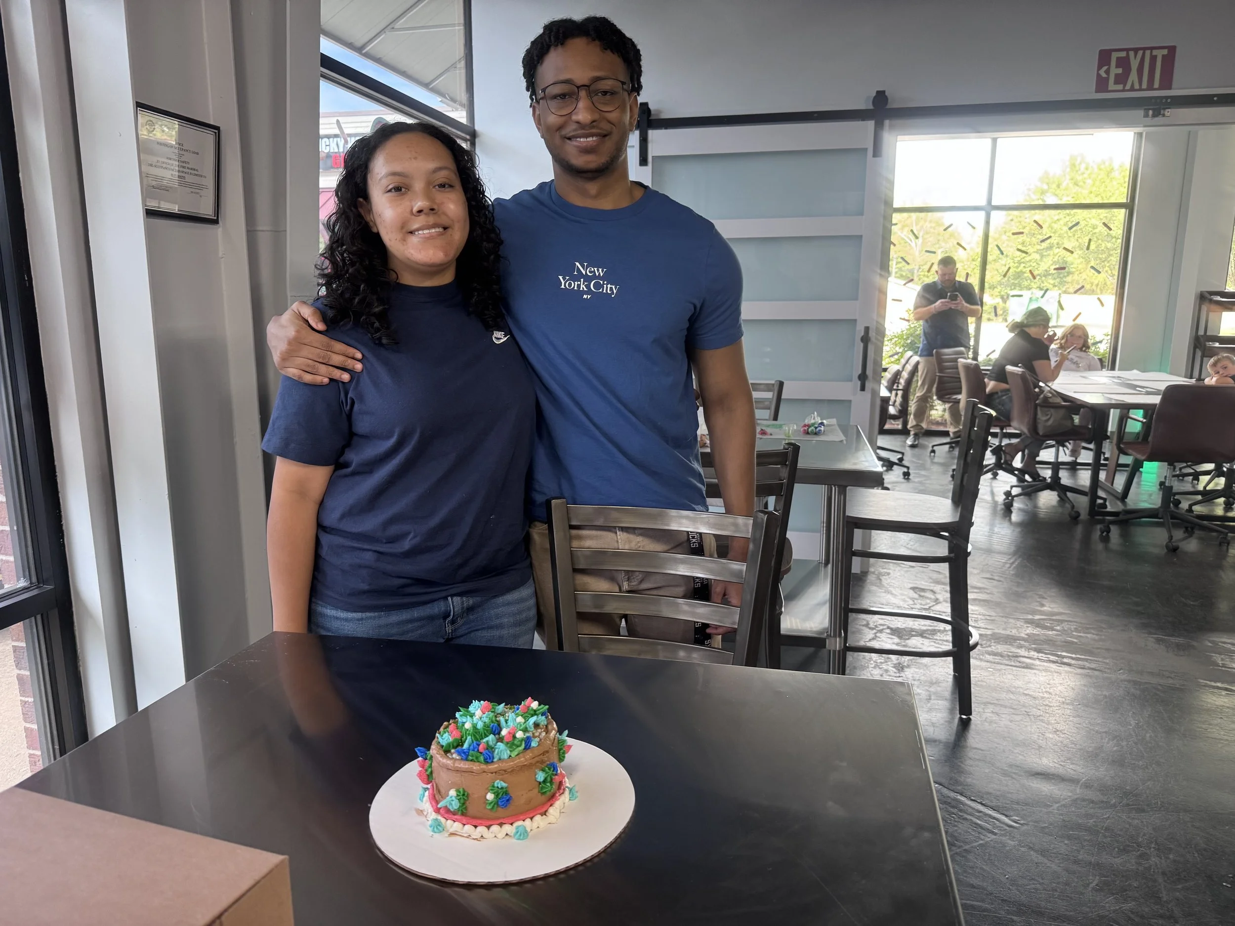 Two people smiling, one woman and one man, standing behind a decorated small cake with colorful frosting and sprinkles on a black table in a bright room with large windows, and a group of people in the background.