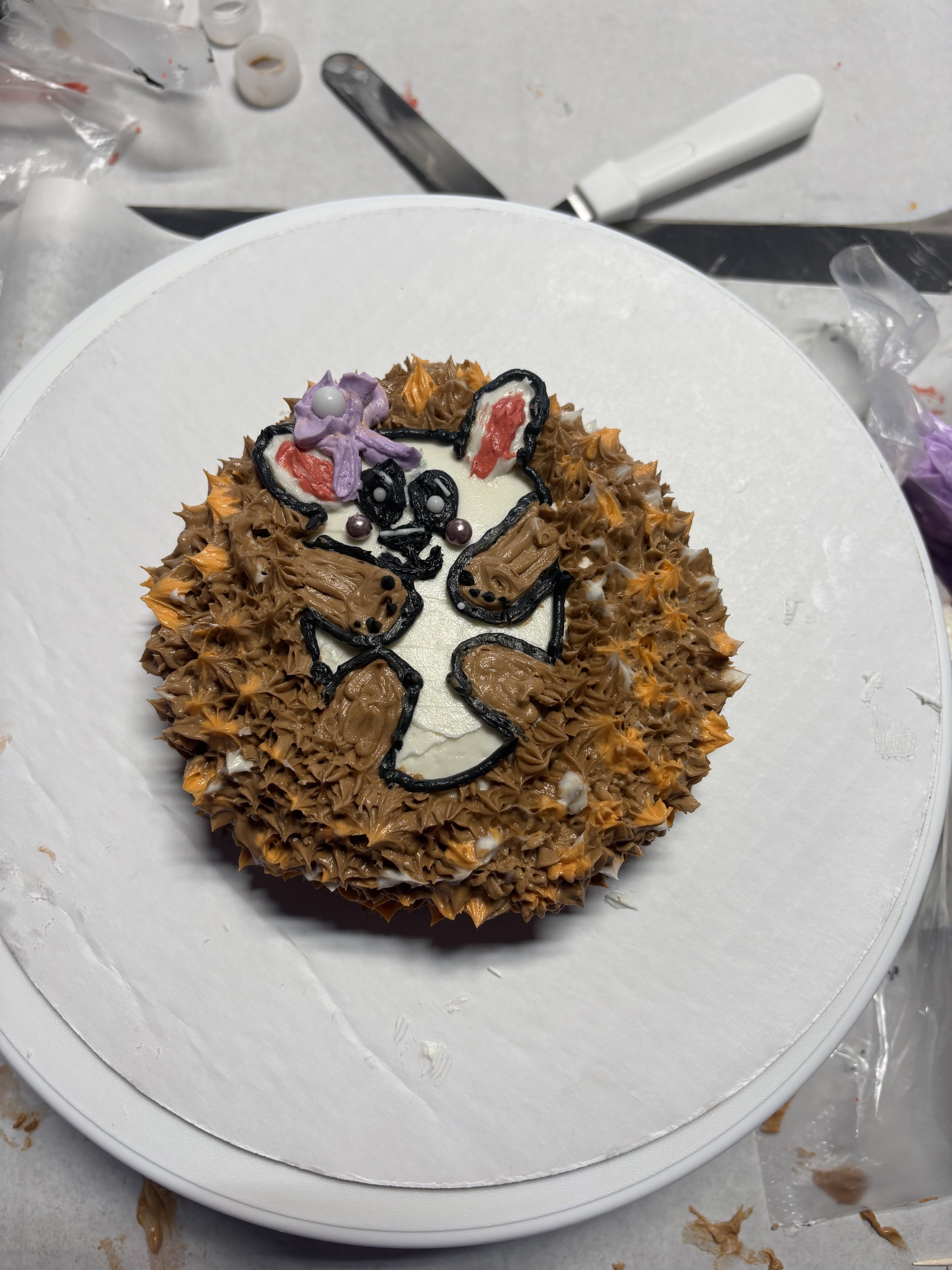 A cake decorated with a hedgehog face made from piped chocolate frosting with purple and white icing details, on a white cake board surrounded by baking tools and ingredients.