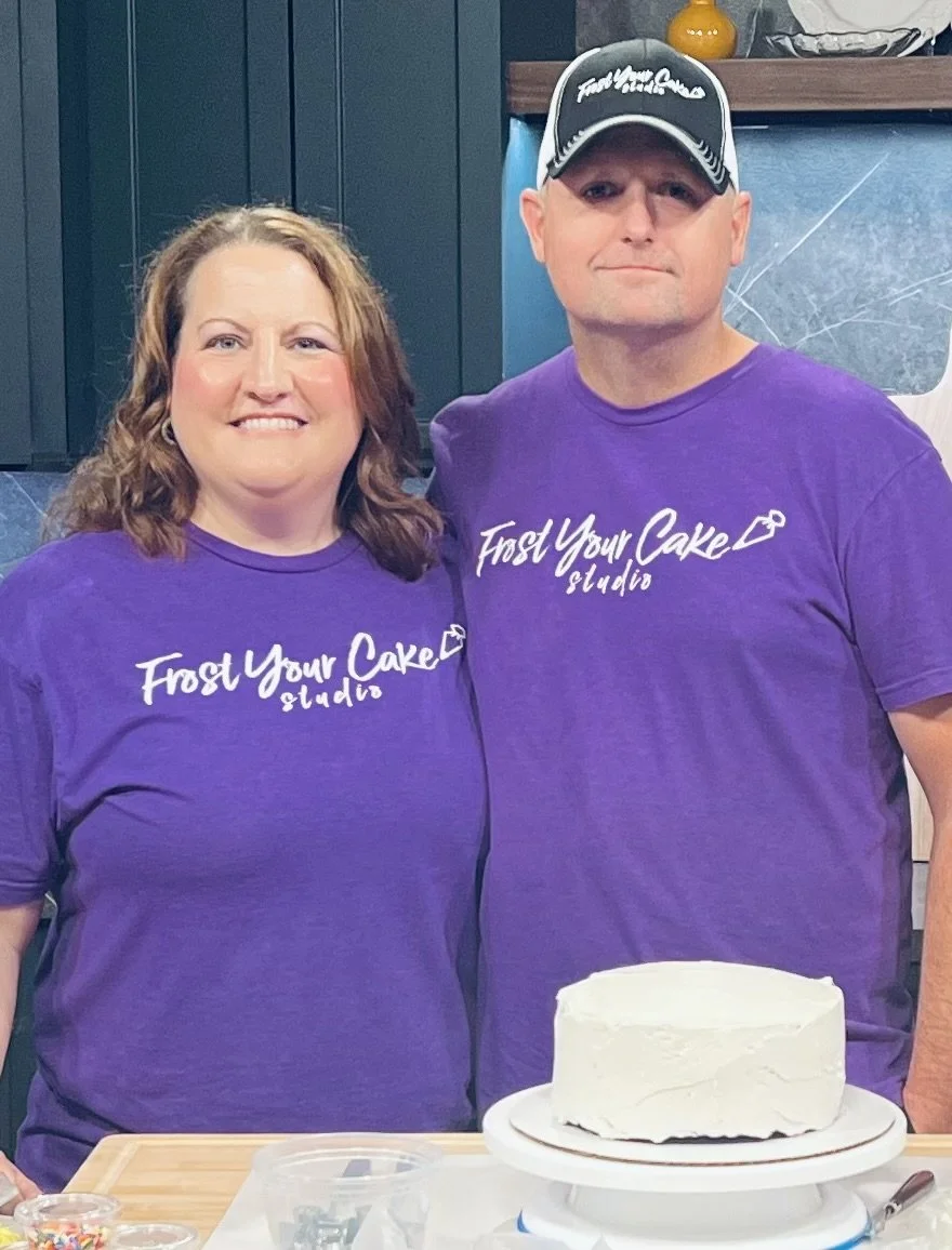 A woman and a man wearing matching purple T-shirts that say "Frost Your Cake studio" stand together in a kitchen, smiling. A white frosted cake on a cake stand is on the counter in front of them.