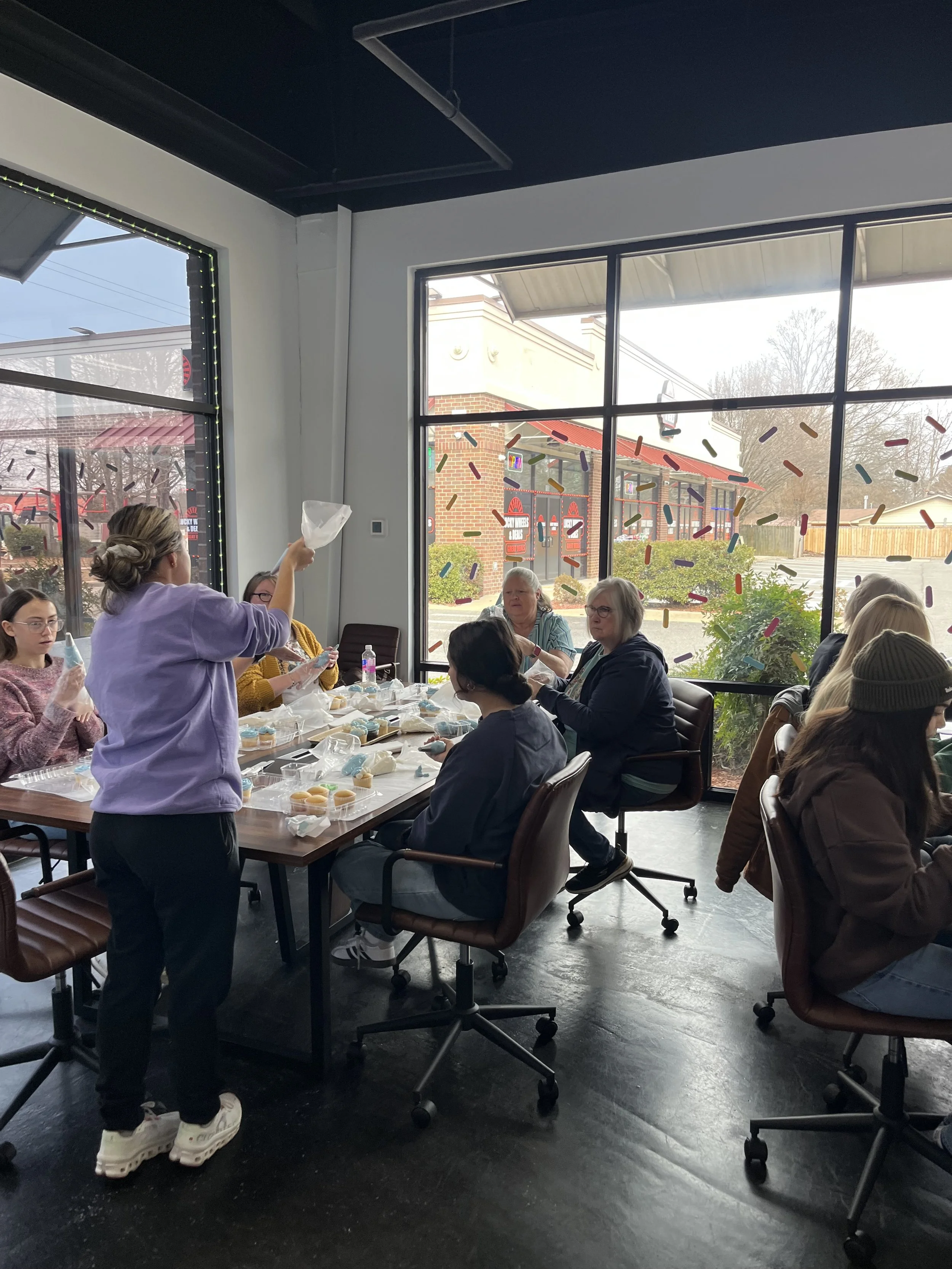People sitting at a table inside a restaurant or cafe, with one person in a lavender shirt standing and holding a plastic bag, others engaging in conversation, large windows showing a building and trees outside, colorful decorations on the windows.