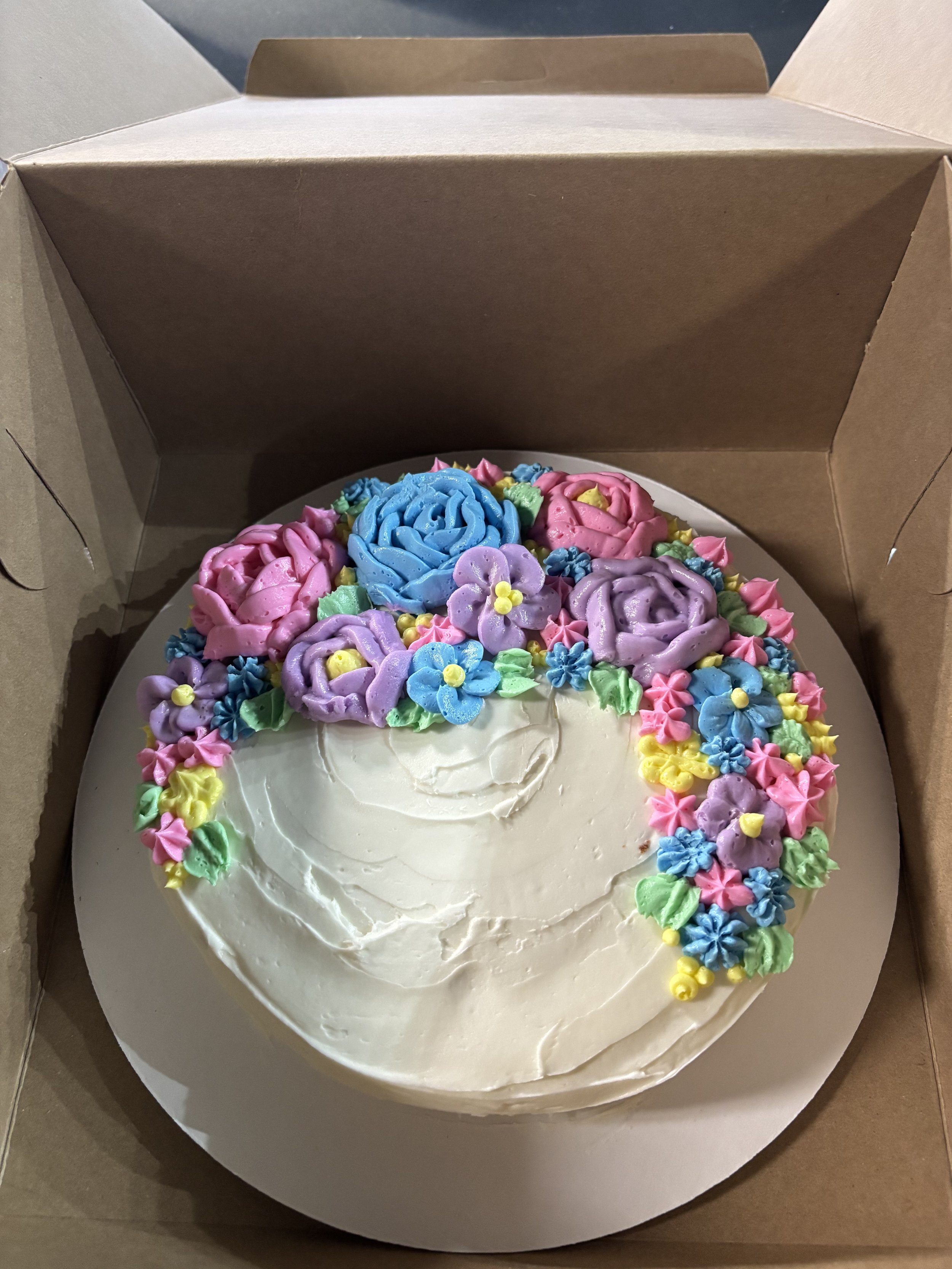 A round cake decorated with colorful buttercream flowers and rosettes, inside a white box with a partially opened brown cardboard lid.