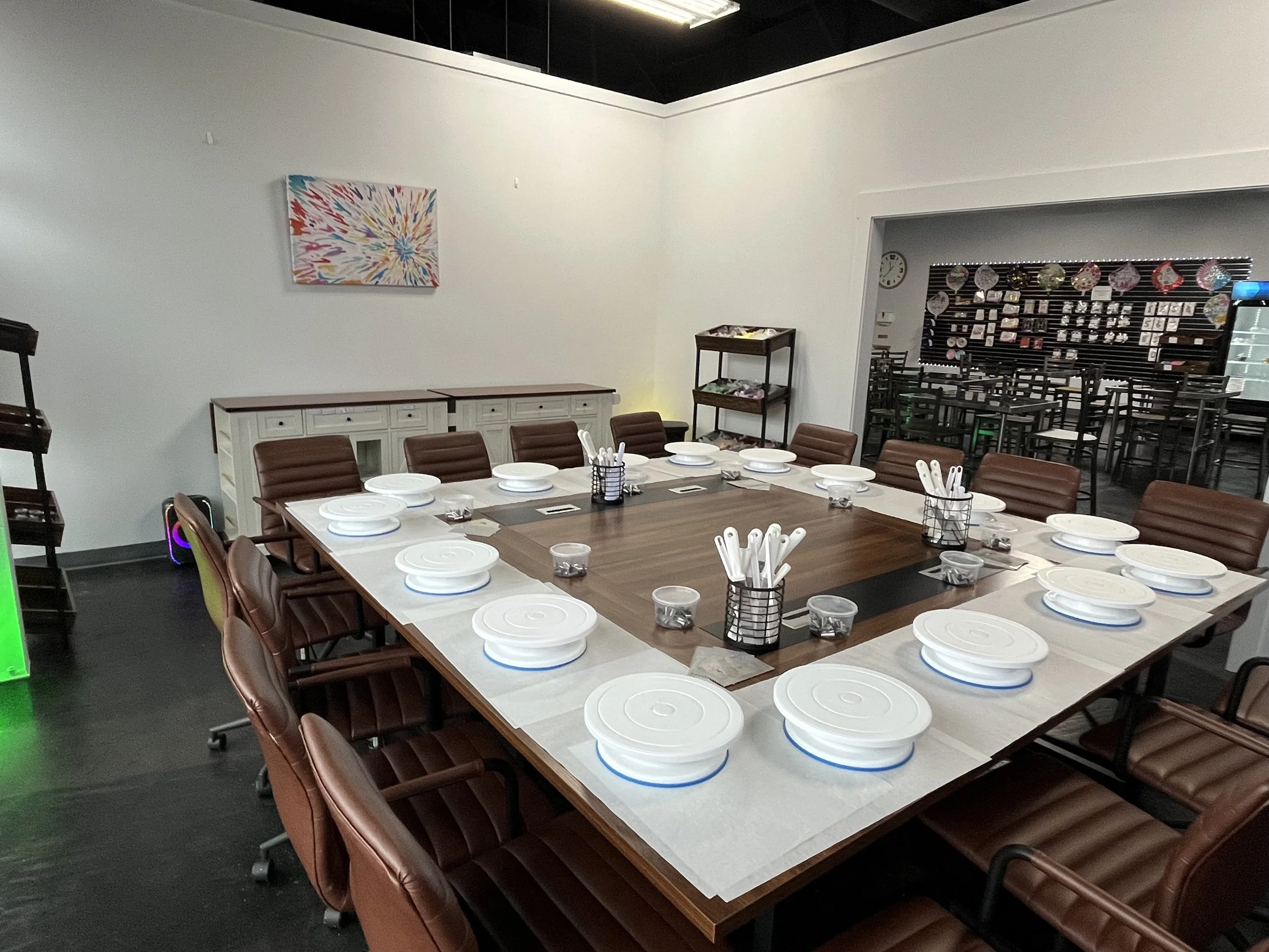 A conference room with a large wooden table set with white plates, utensils, and small cups, surrounded by brown chairs. In the background, there are white cabinets and a small display rack. The walls are decorated with colorful artwork, and another room can be seen through an open doorway, featuring black chairs and a wall display with hanging decorations.