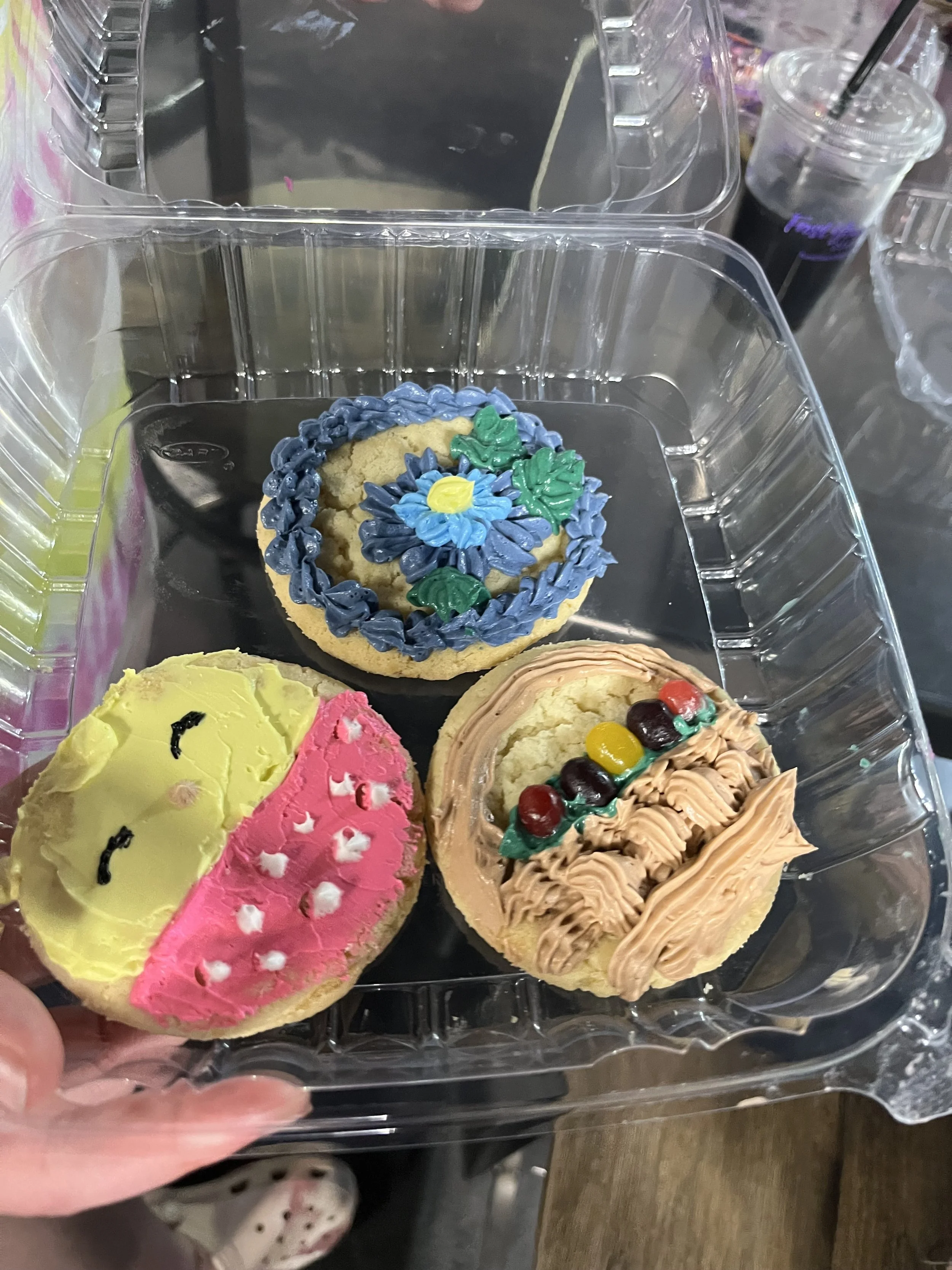 Assorted decorated cookies in a clear plastic container, with a hand holding the container. The cookies are decorated with colorful frosting and toppings.