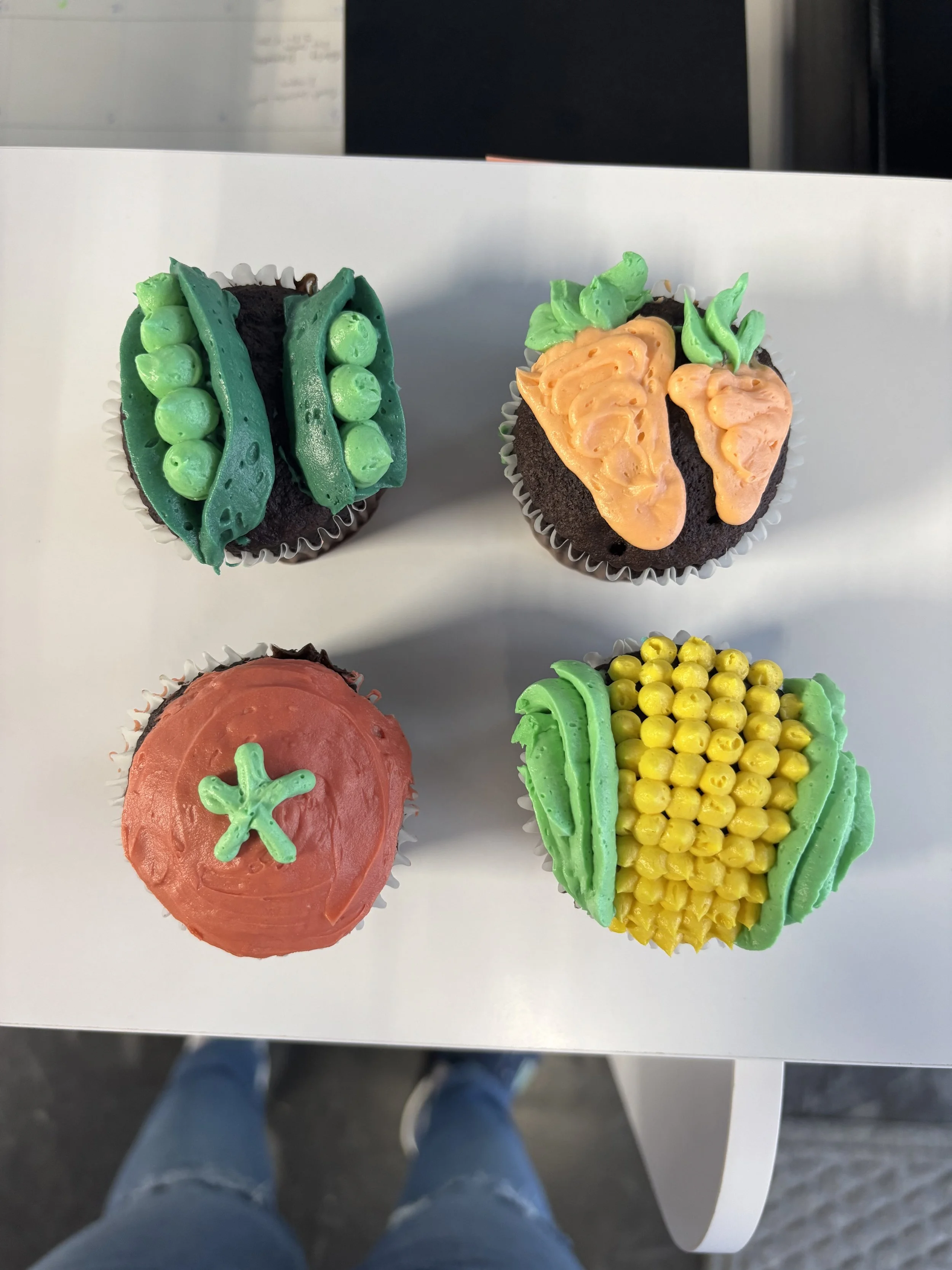 Decorated cupcakes with colorful frosting in the shape of vegetables, including peas, carrots, and corn, topped with icing and edible decorations, displayed on a white surface.