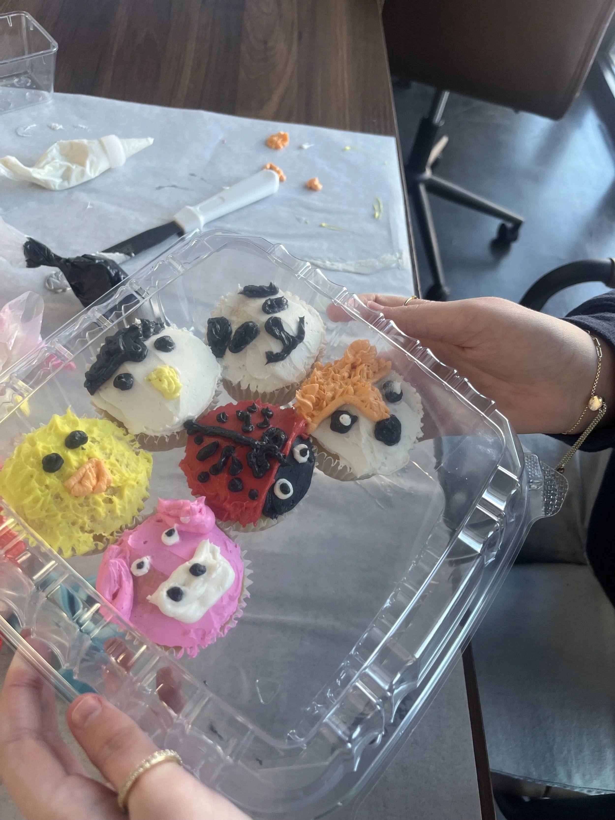 A hand holding a clear plastic container with decorated cupcakes featuring animal faces and colorful frosting, on a table with baking tools and leftover decorations.