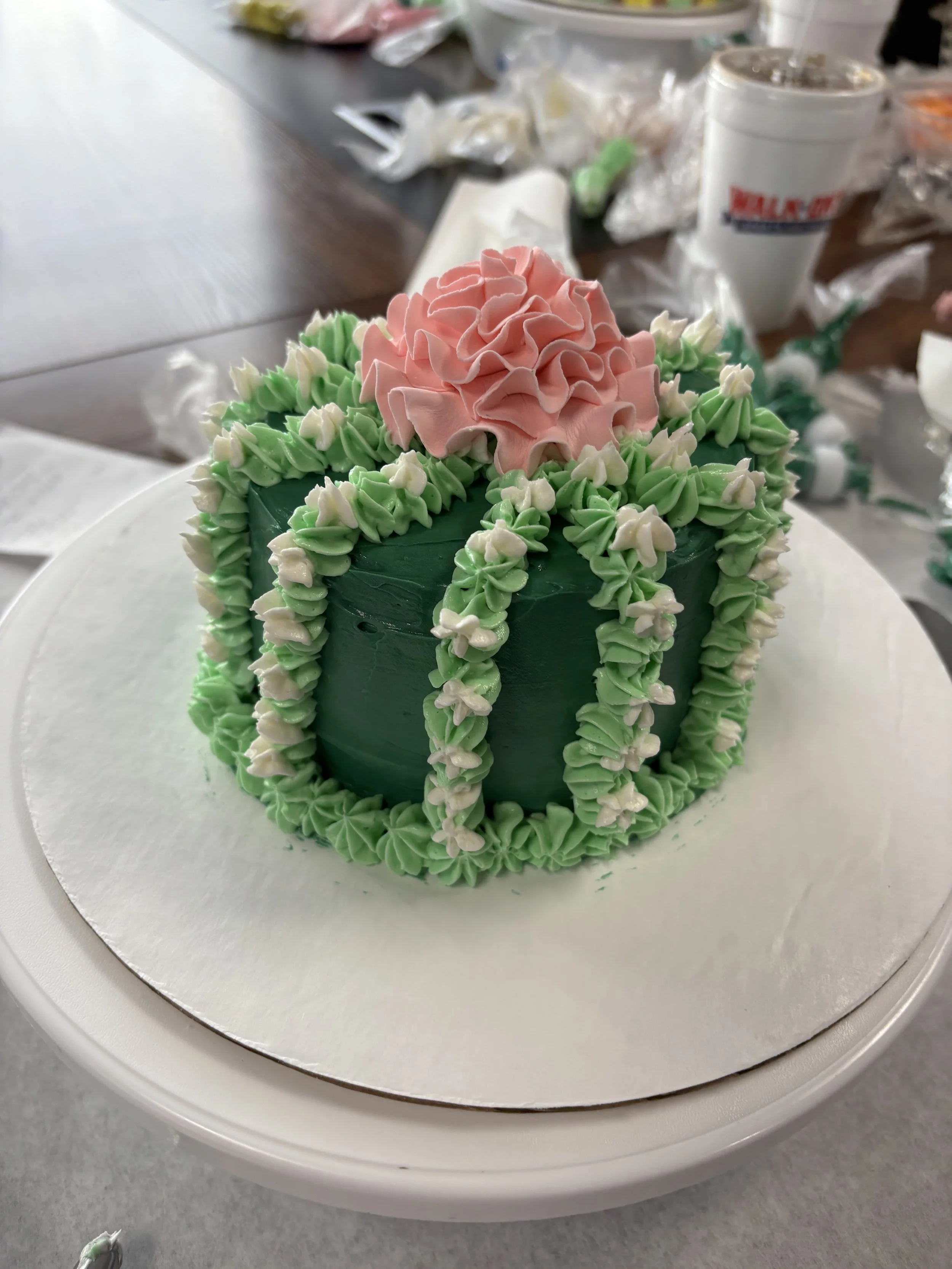 Decorative cake with pink flower on top, green and white piped frosting decorations, on a white cake board.