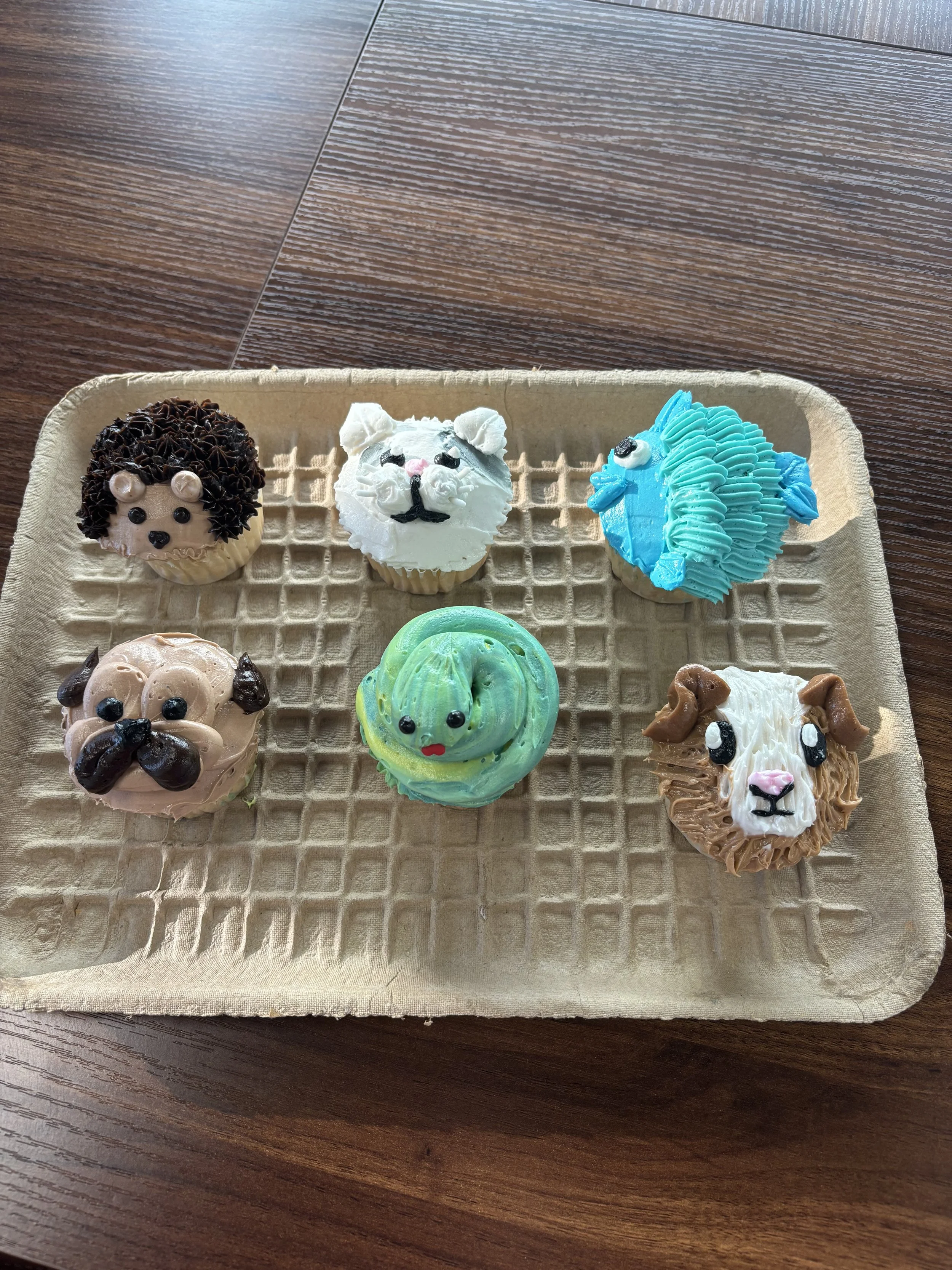 Assorted animal-themed cupcakes on a tray, including a dog, a fox, a horse, a panda, a turtle, and a dog, decorated with colorful frosting and edible details.
