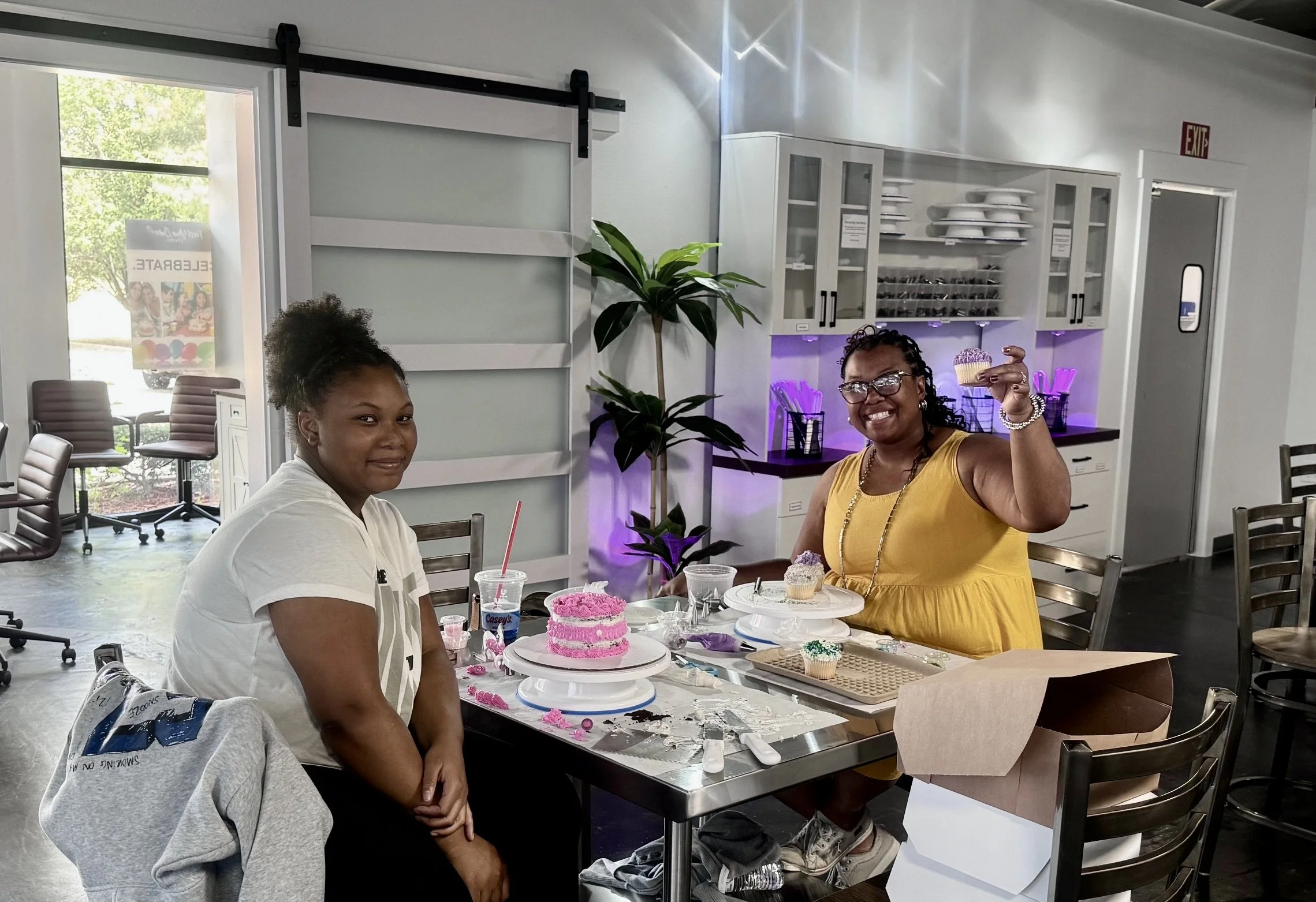 Two women celebrating a birthday at a restaurant, with pink birthday cake and cupcakes on the table, one woman is smiling and holding a cupcake, the other is sitting and smiling, in a brightly lit restaurant with white cabinets, a plant, and soccer chairs.