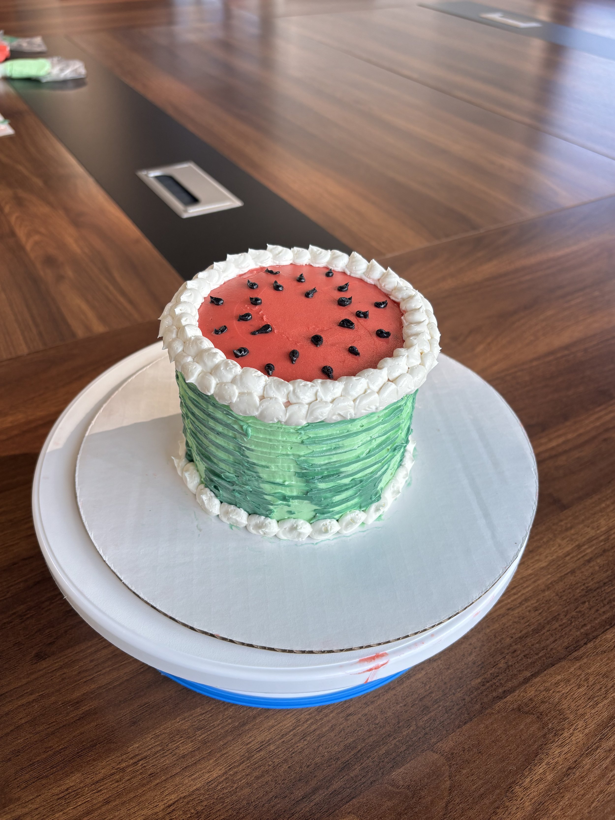 A watermelon-shaped cake with white and green icing, topped with black seeds, sitting on a round white cake board on a dark wooden table.