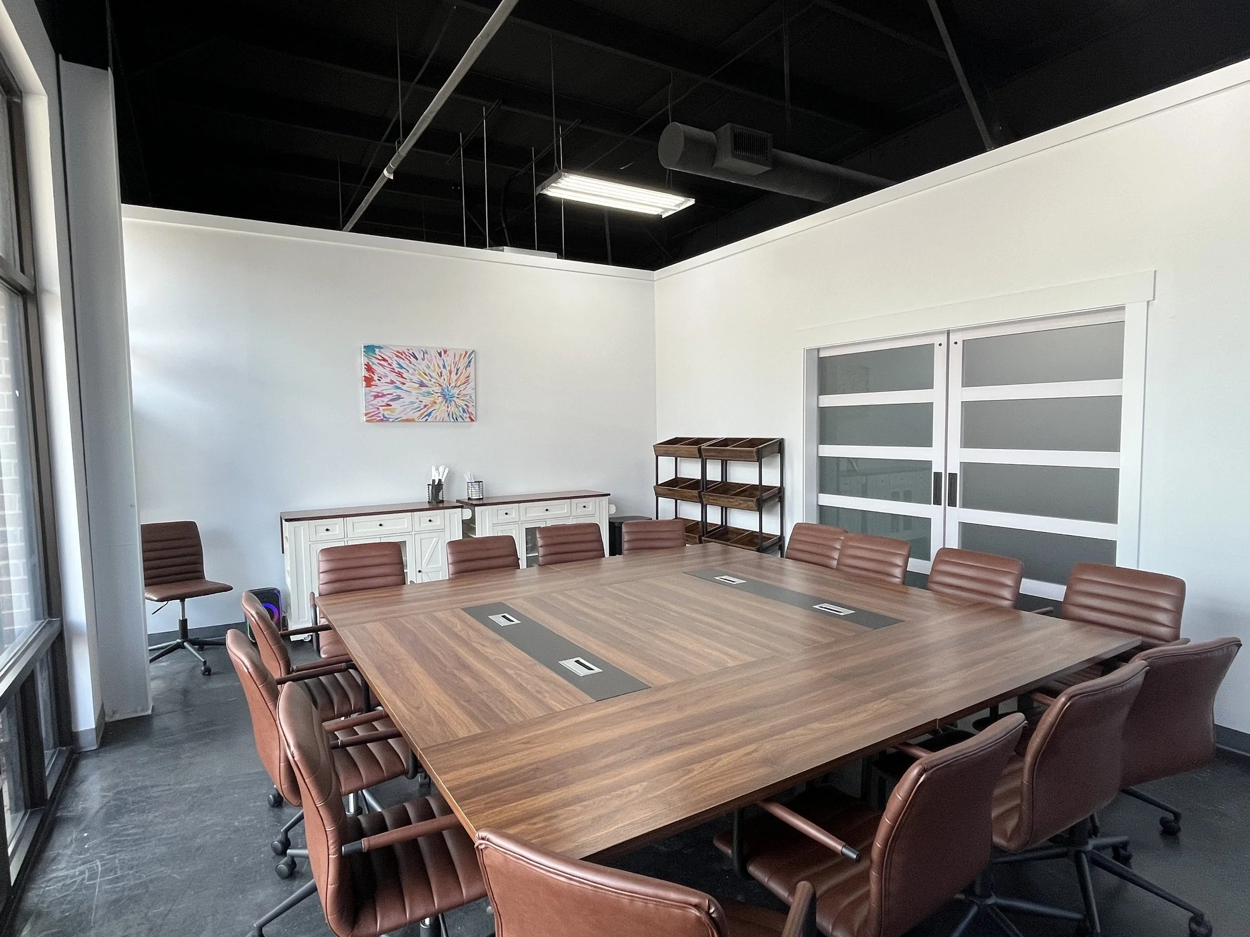 A conference room with a long wooden table surrounded by brown chairs. The room has white walls, a white cabinet, a colorful modern art piece on the wall, and a black ceiling with lighting and air ducts.