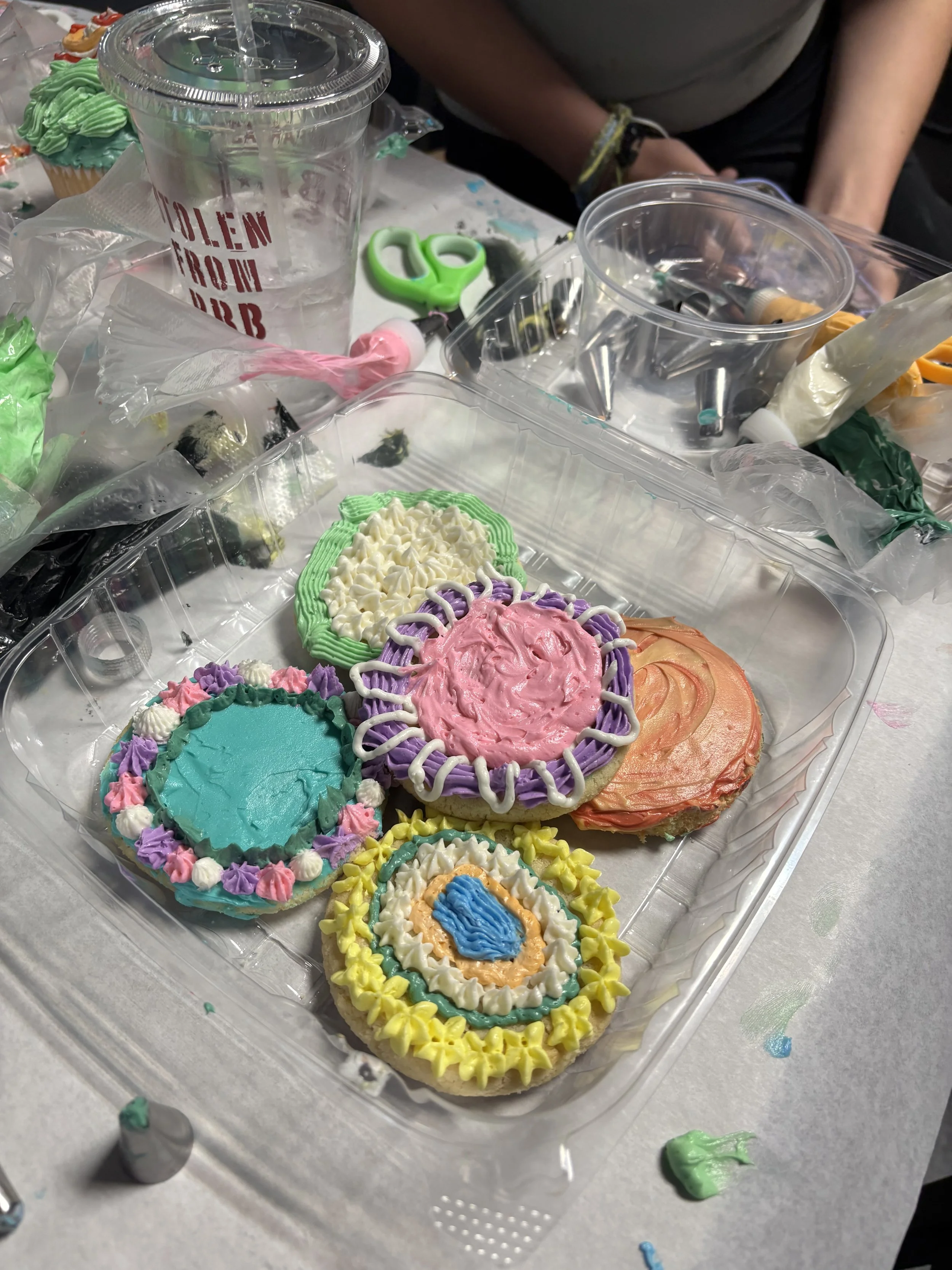 Colorfully decorated cookies with various icing designs on a clear plastic tray, surrounded by decorating tools like piping bags, scissors, and a cup of water on a white table.