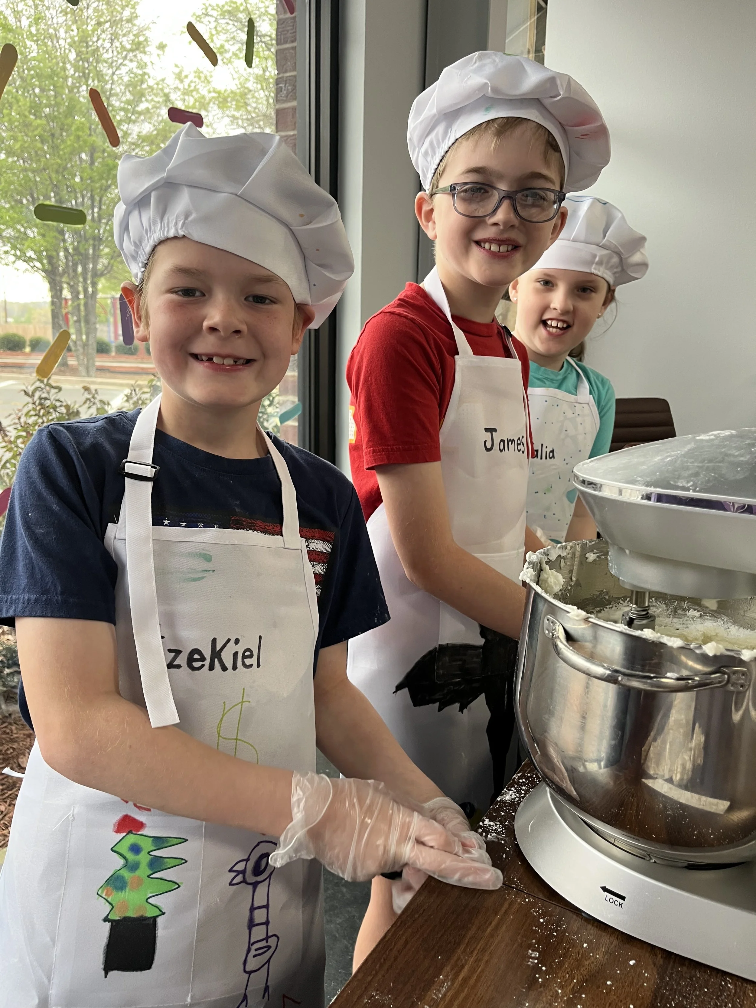 Three children wearing chef hats and aprons are making frosting at a junior decorator spring break camp.