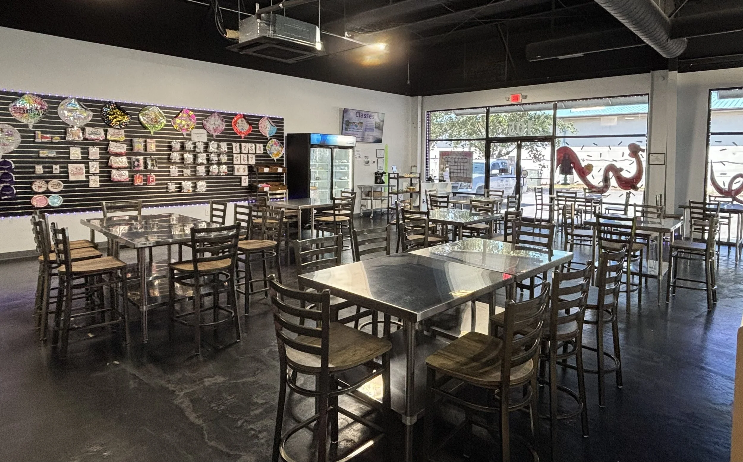 Empty restaurant with metal tables and wooden chairs, decorated wall with colorful balloons and other decorations, large windows showing outside parking lot and trees, Pepsi refrigerator, and digital menu display.