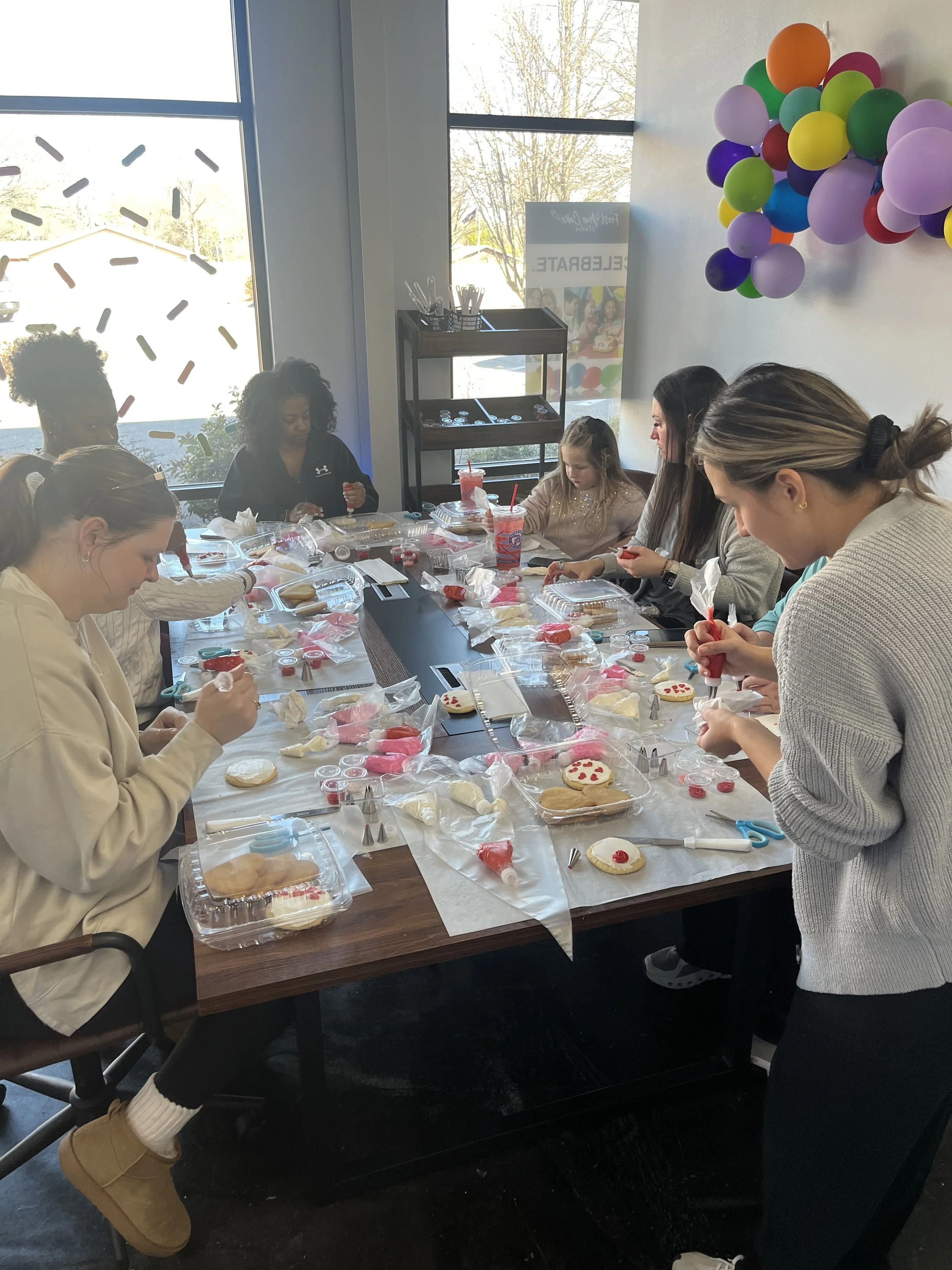 Group of people, including children and adults, decorating cookies with icing at a table during a celebration. There are balloons on the wall, and the table is filled with cookies, icing, and baking supplies.