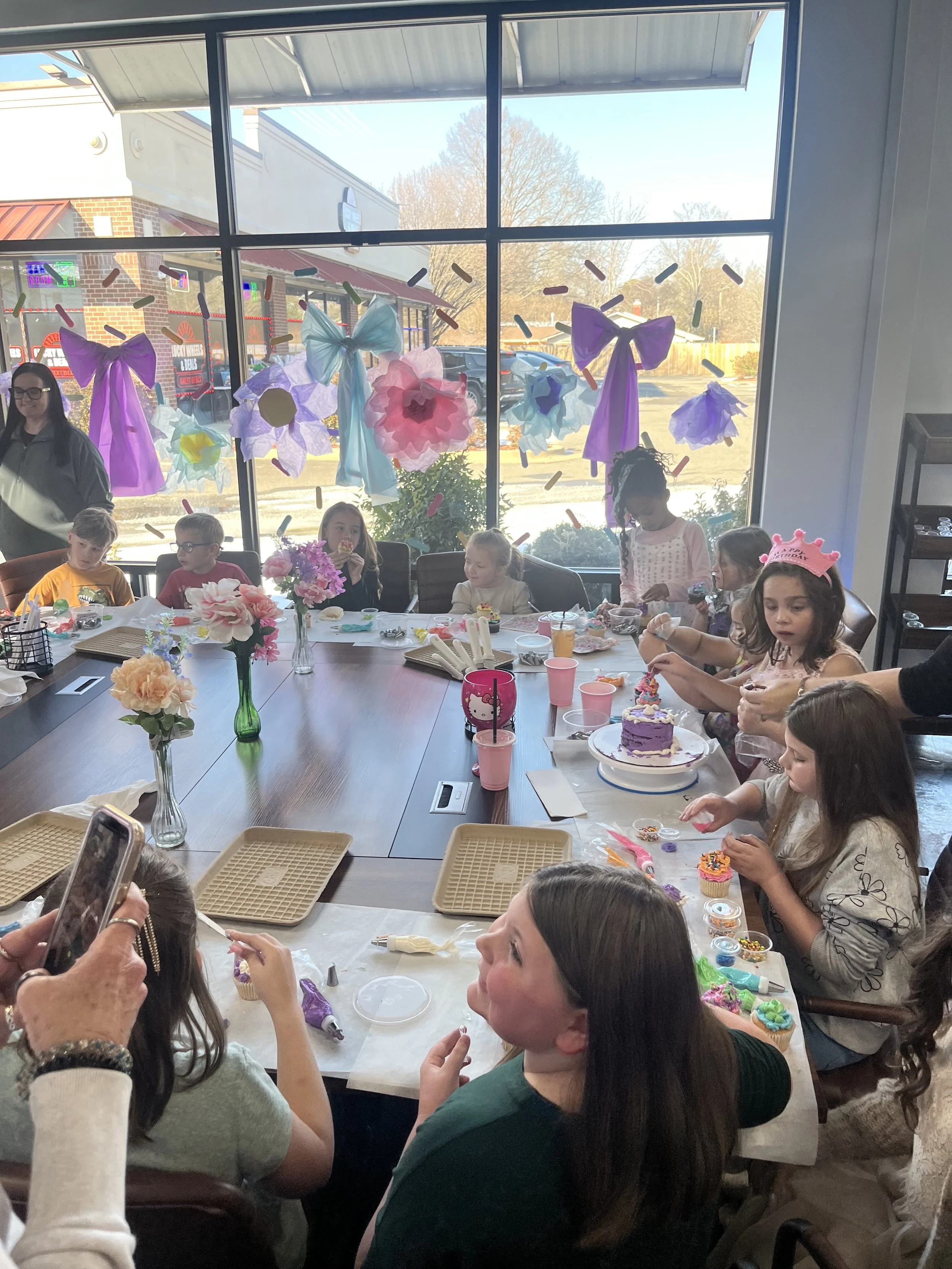 Children and adults celebrating a birthday party at a restaurant or cafe. The table is decorated with flowers and cupcakes, and there are birthday hats and party decorations, including large purple and blue bows on the window.