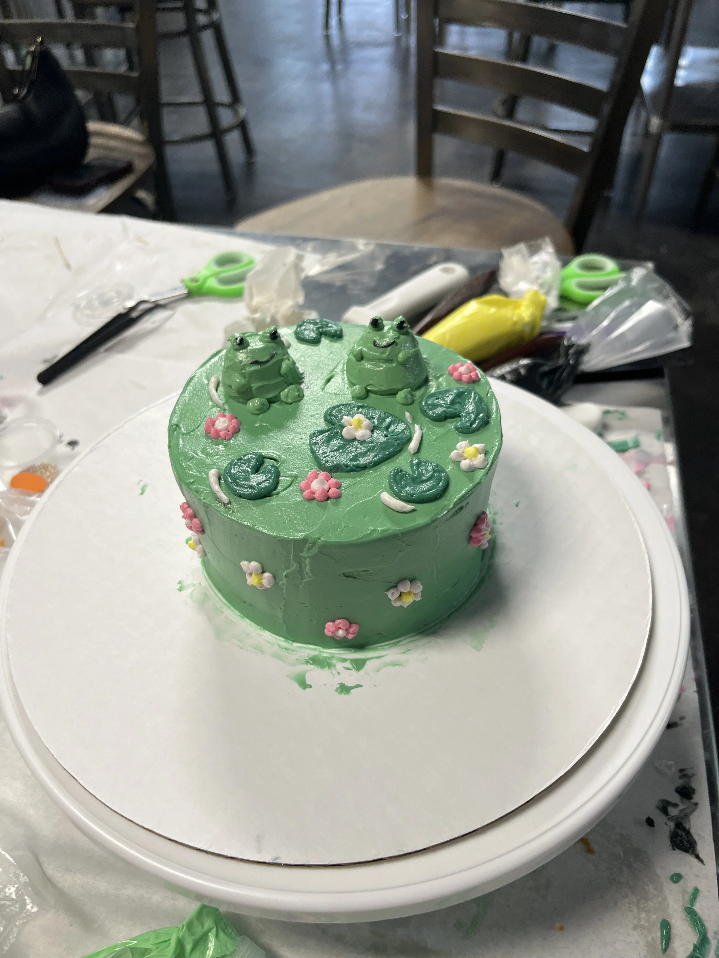 A round, green cake decorated with frogs, flowers, and lily pads, placed on a white surface with some decorating tools around it.