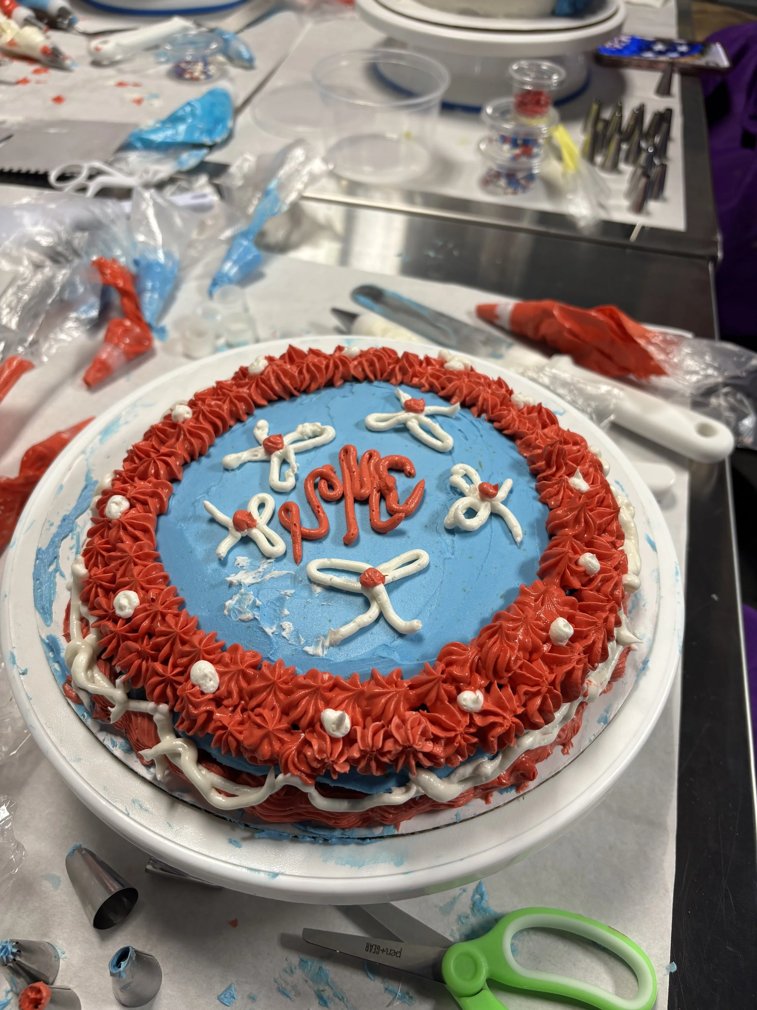 Decorated birthday cake with blue, red, and white frosting, with the words 'Happy Birthday' written on top amid celebratory icing decorations, placed on a table with baking tools and supplies.