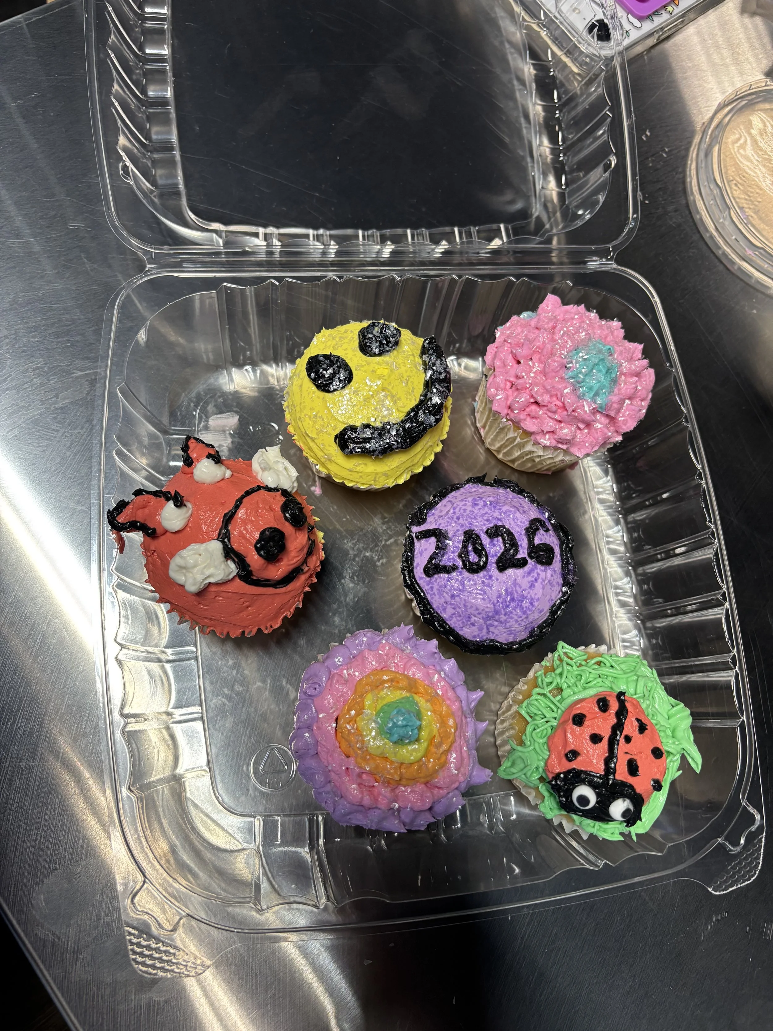 Assorted colorful cupcakes decorated with frosting, sprinkles, and fondant in a clear plastic container.