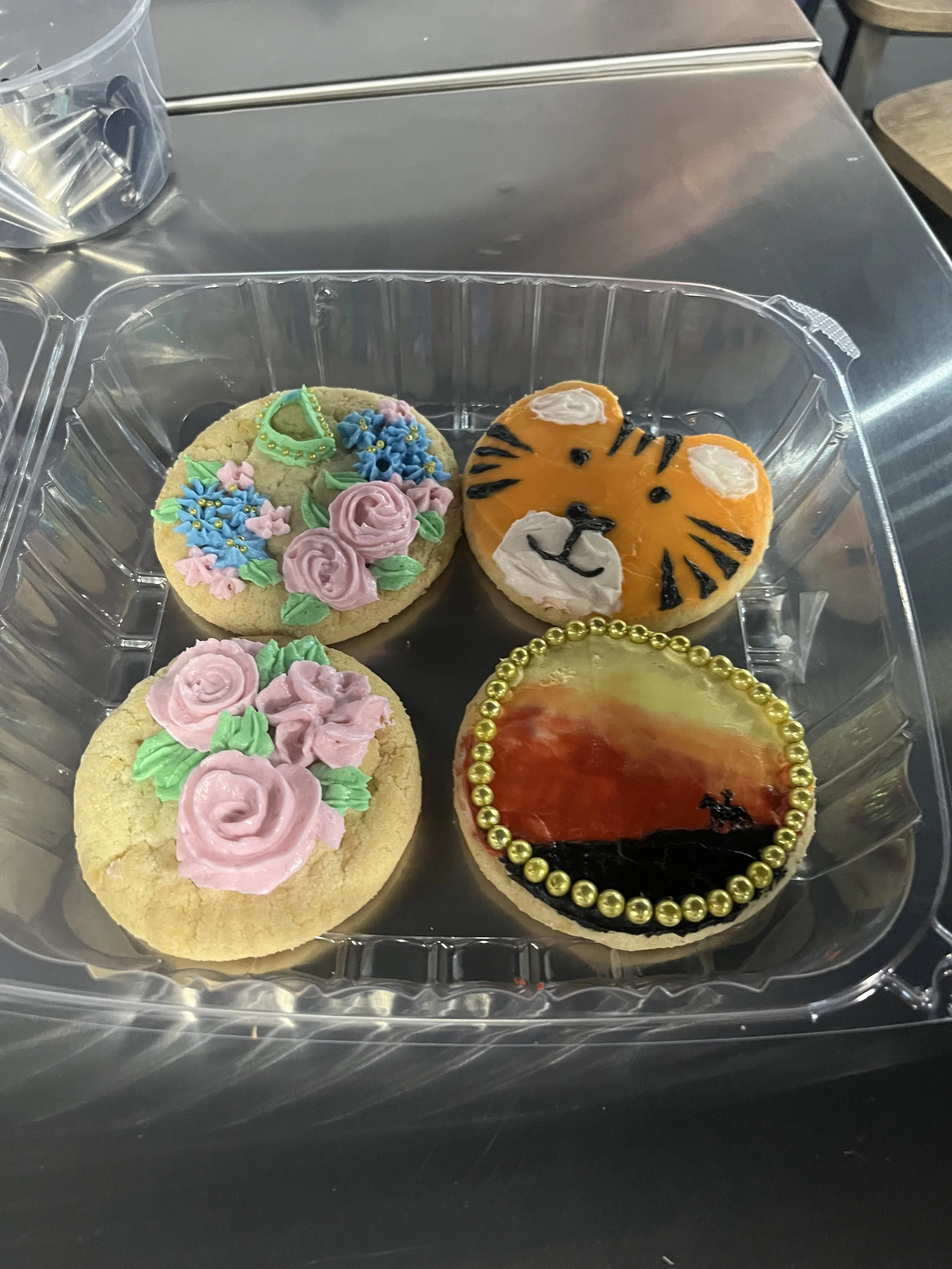 Decorated cookies inside a plastic container, one shaped like a tiger's face with orange icing and black details, another with pink roses and blue flowers, and part of a third cookie with pink icing roses and green leaves.