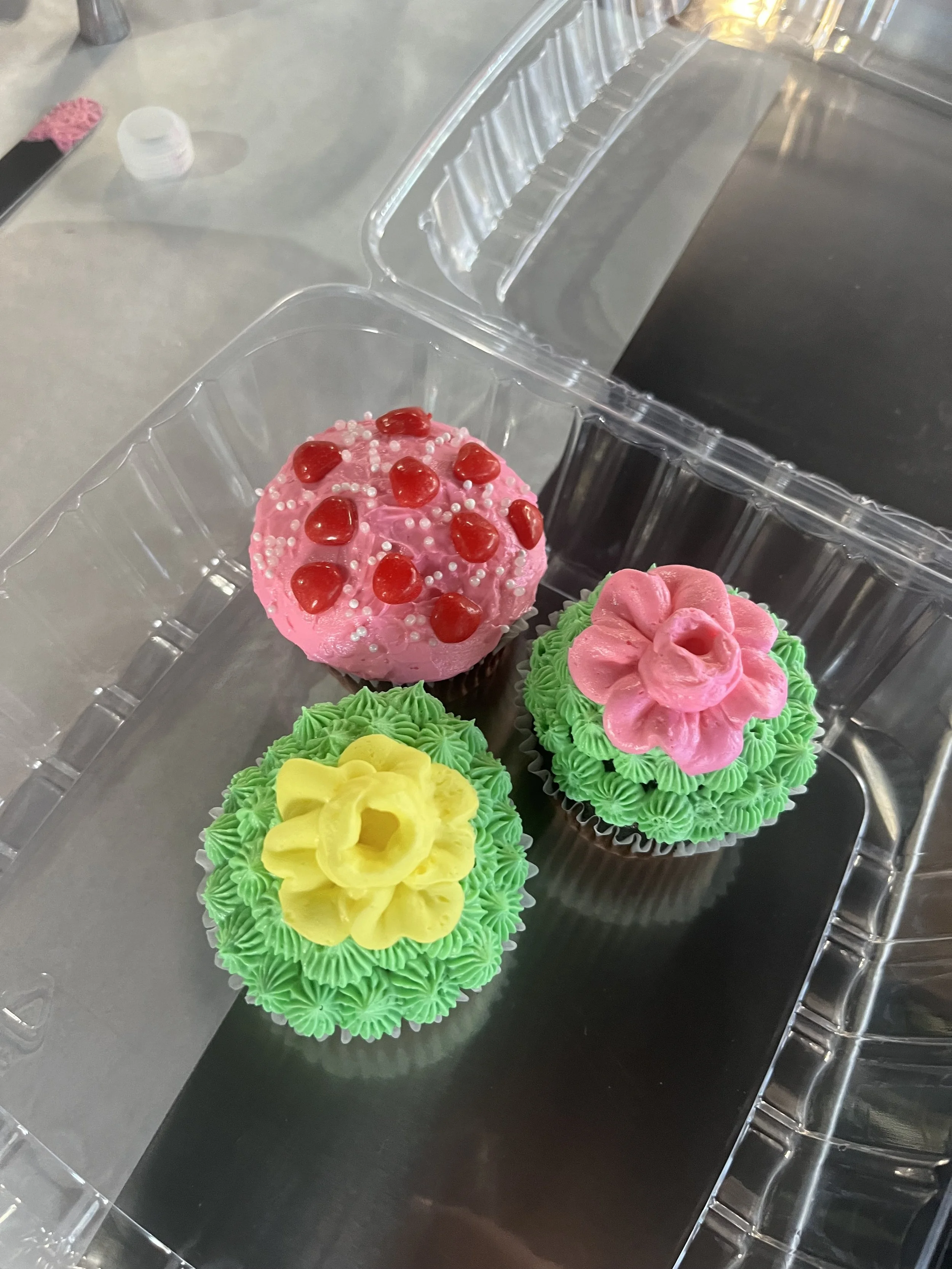 Four decorated cupcakes inside a clear plastic container on a black surface, with a pink one featuring red fruit and white sprinkles, a pink one with pink frosting and a flower shape, and two yellow cupcakes with green frosting and yellow flower decorations.