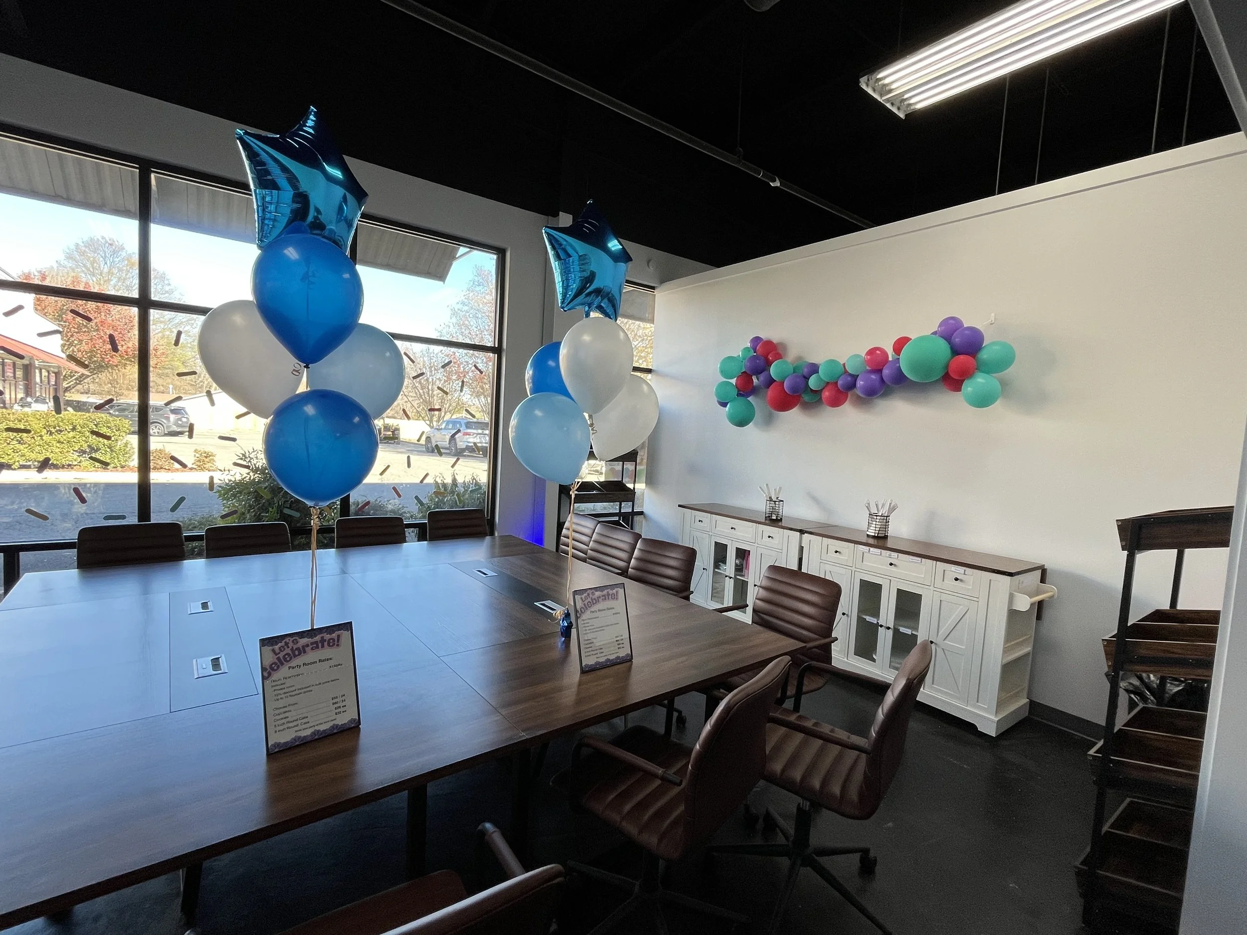 Decorated room with balloons in blue, white, purple, and red colors, set up for a celebration, with a long wooden table and chairs, large windows with outside views, and colorful confetti cutouts on the window glass.