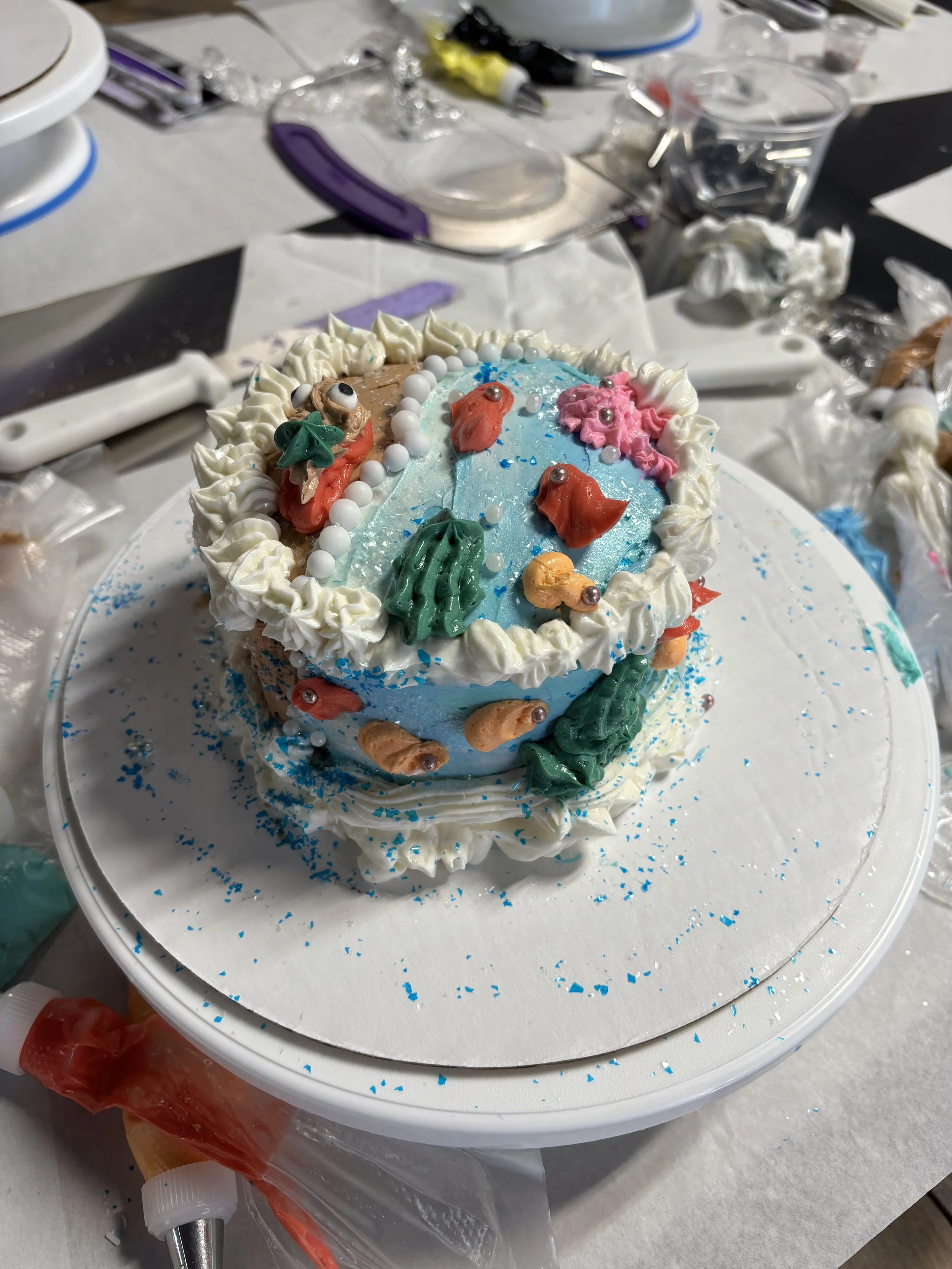 Decorated cake with white frosting, colorful fondant decorations of fish, seaweed, and bubbles, on a white circular cake board.
