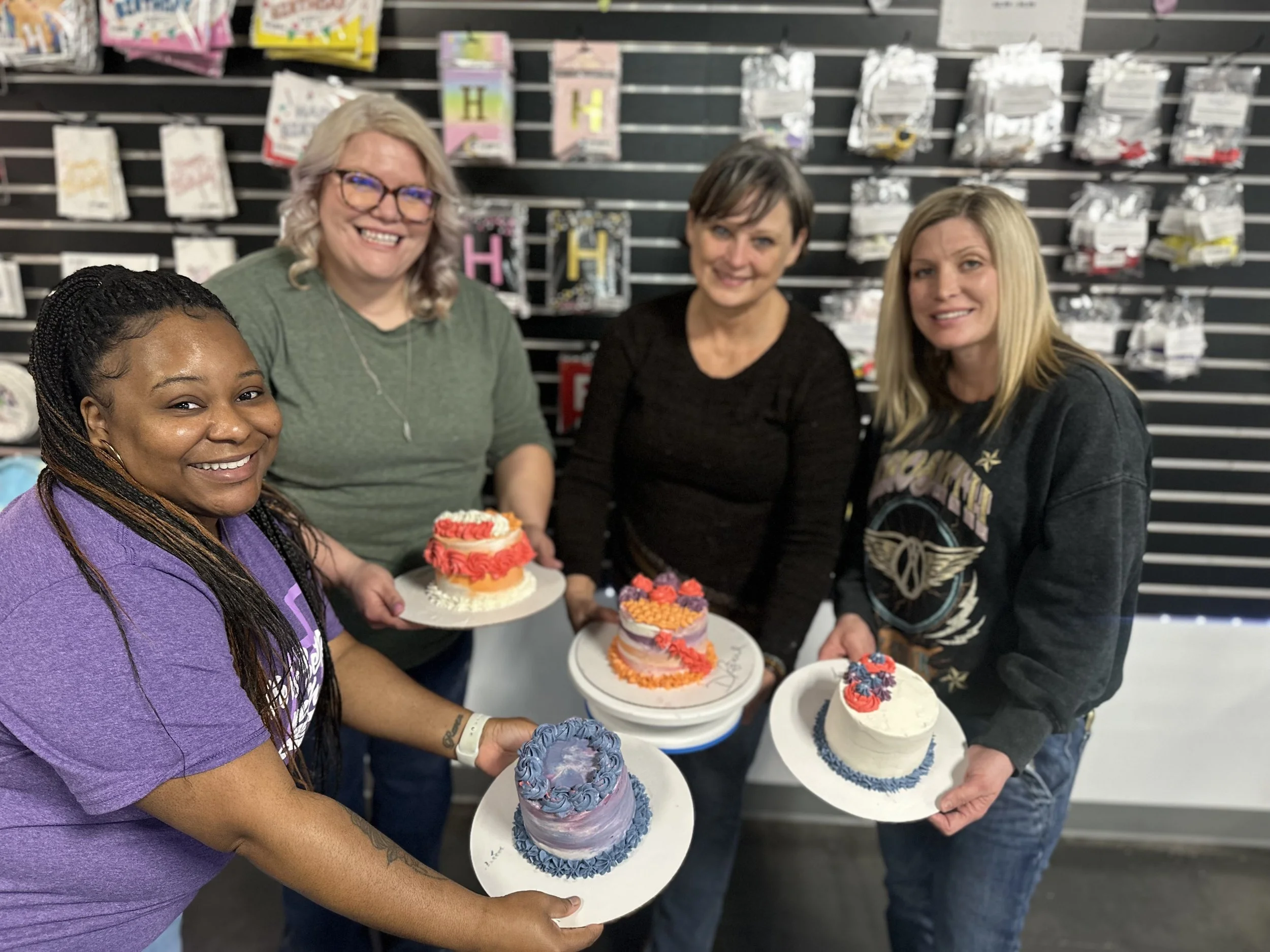 Four women standing in front of a black retail wall, holding cakes decorated during a private decorating class with colorful frosting and designs, smiling at the camera.