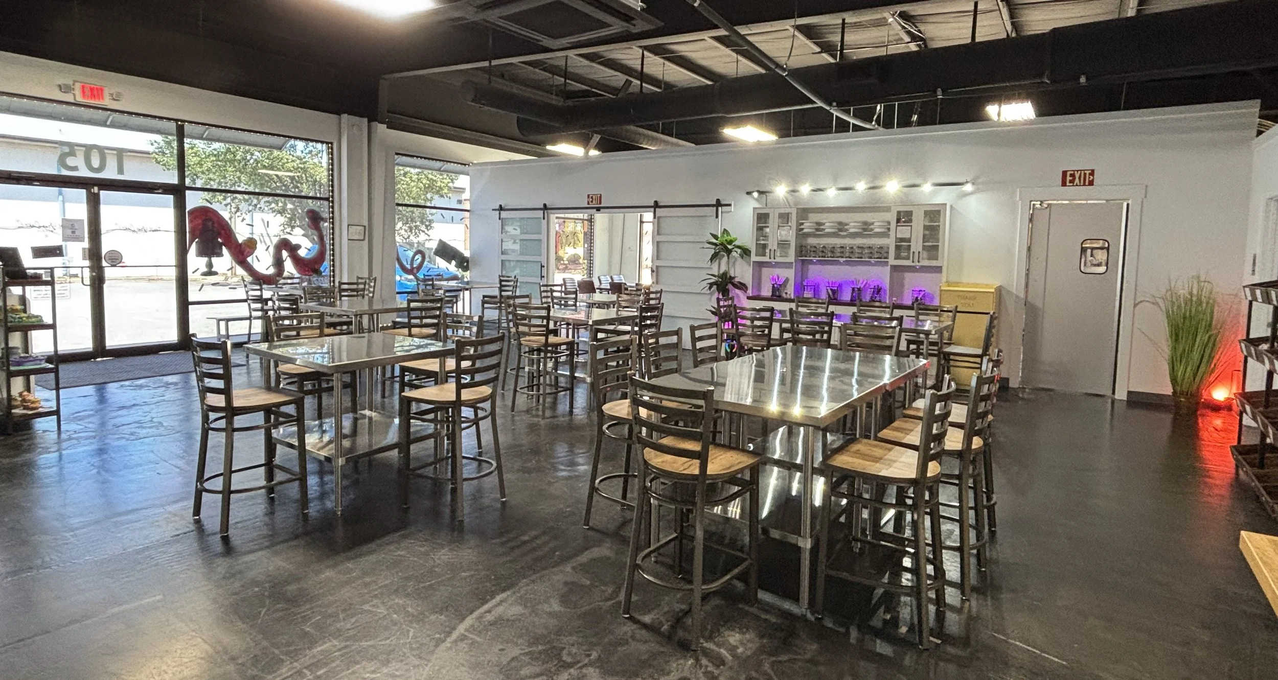 Interior of a modern cafe with multiple tables and chairs, large windows showing the street outside, some decorative plants, and a colorful light display on the wall.