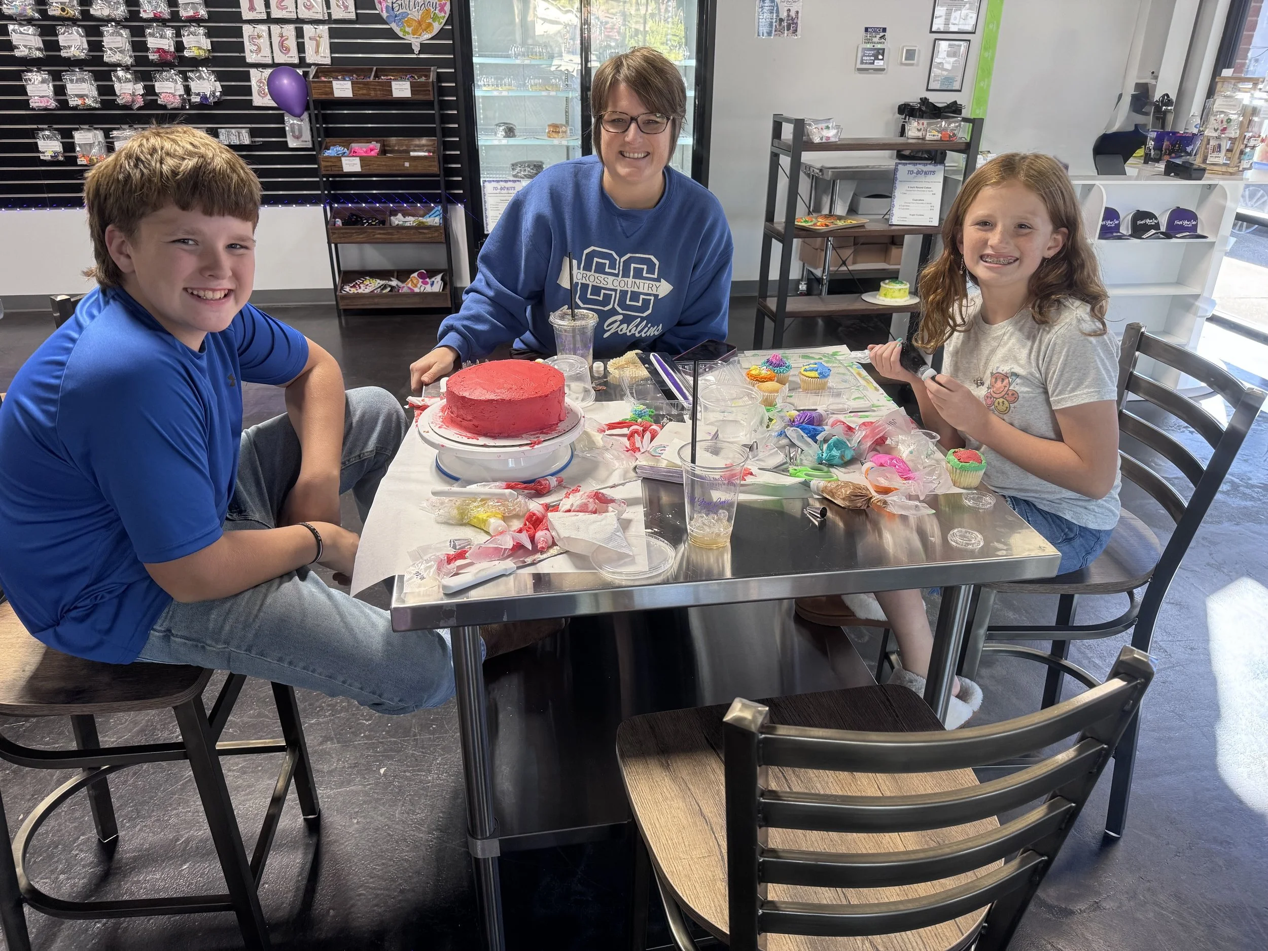 Three kids and one woman sitting at a table decorated with cupcakes and a red-frosted birthday cake. The girl on the right is holding a black object, smiling. The boy on the left and the woman are smiling at the camera.