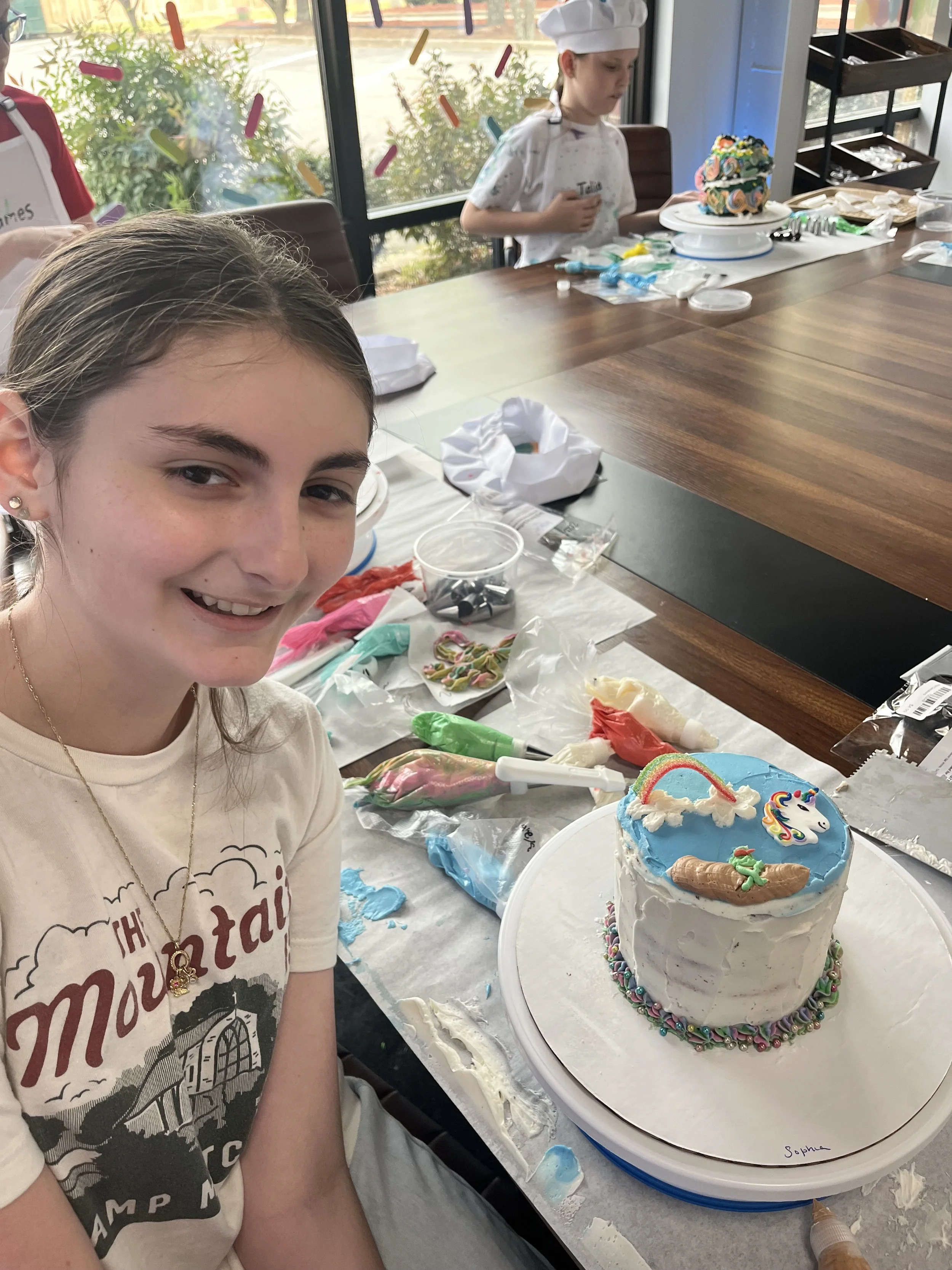 Two young girls are celebrating with cakes in what appears to be a bakery or cake decorating workshop. The girl in the foreground is smiling at the camera, with a partially decorated birthday cake featuring a unicorn, rainbows, and a shamrock on her table. The other girl, in the background, is focused on decorating her cake. The table is filled with various icing tools, colorful decorations, and supplies.