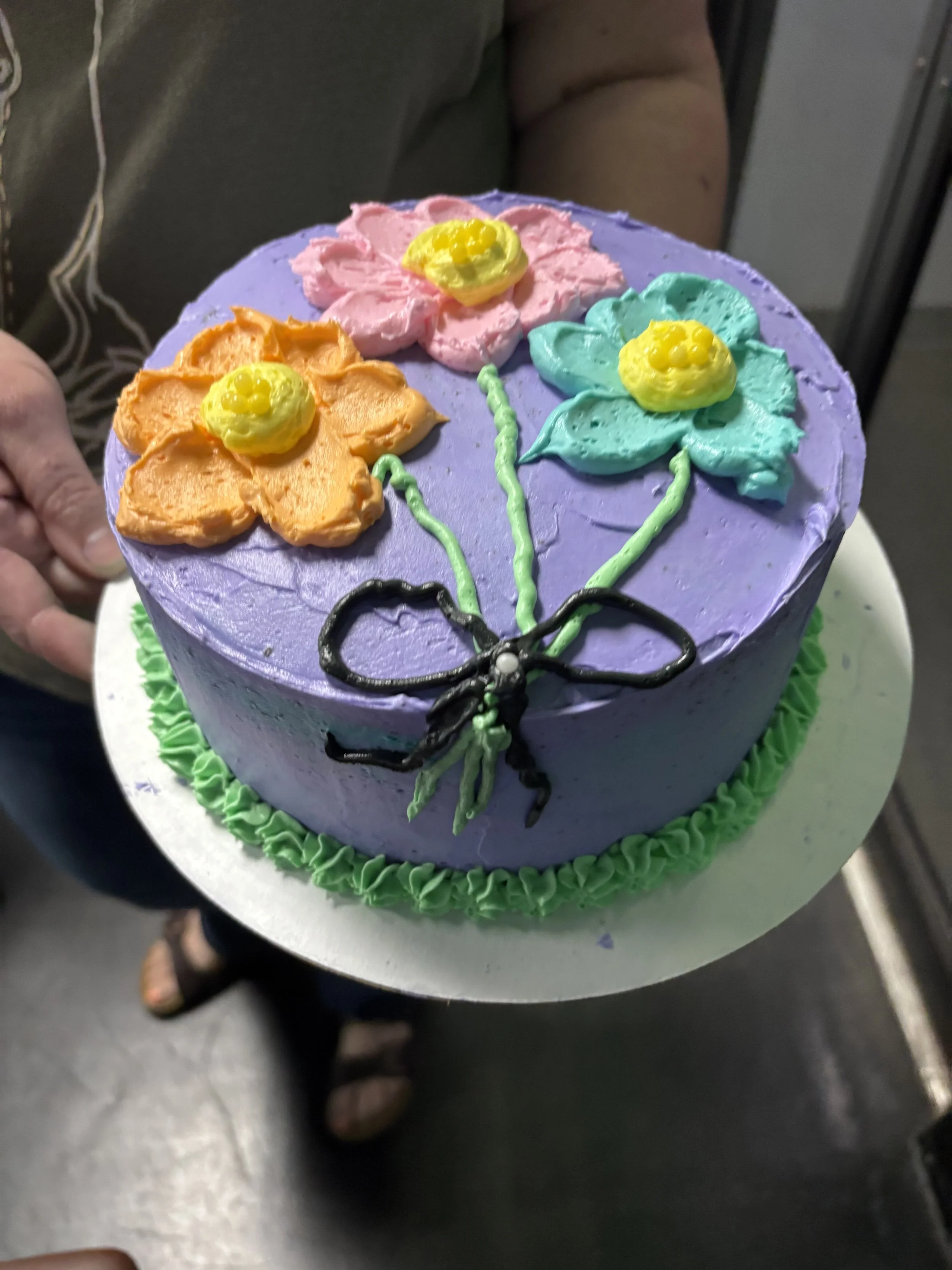 Colorful birthday cake with purple frosting, decorated with three flower-shaped frosting designs in pink, orange, and teal, each with a yellow frosting center, and a black icing bow with green stems at the base.