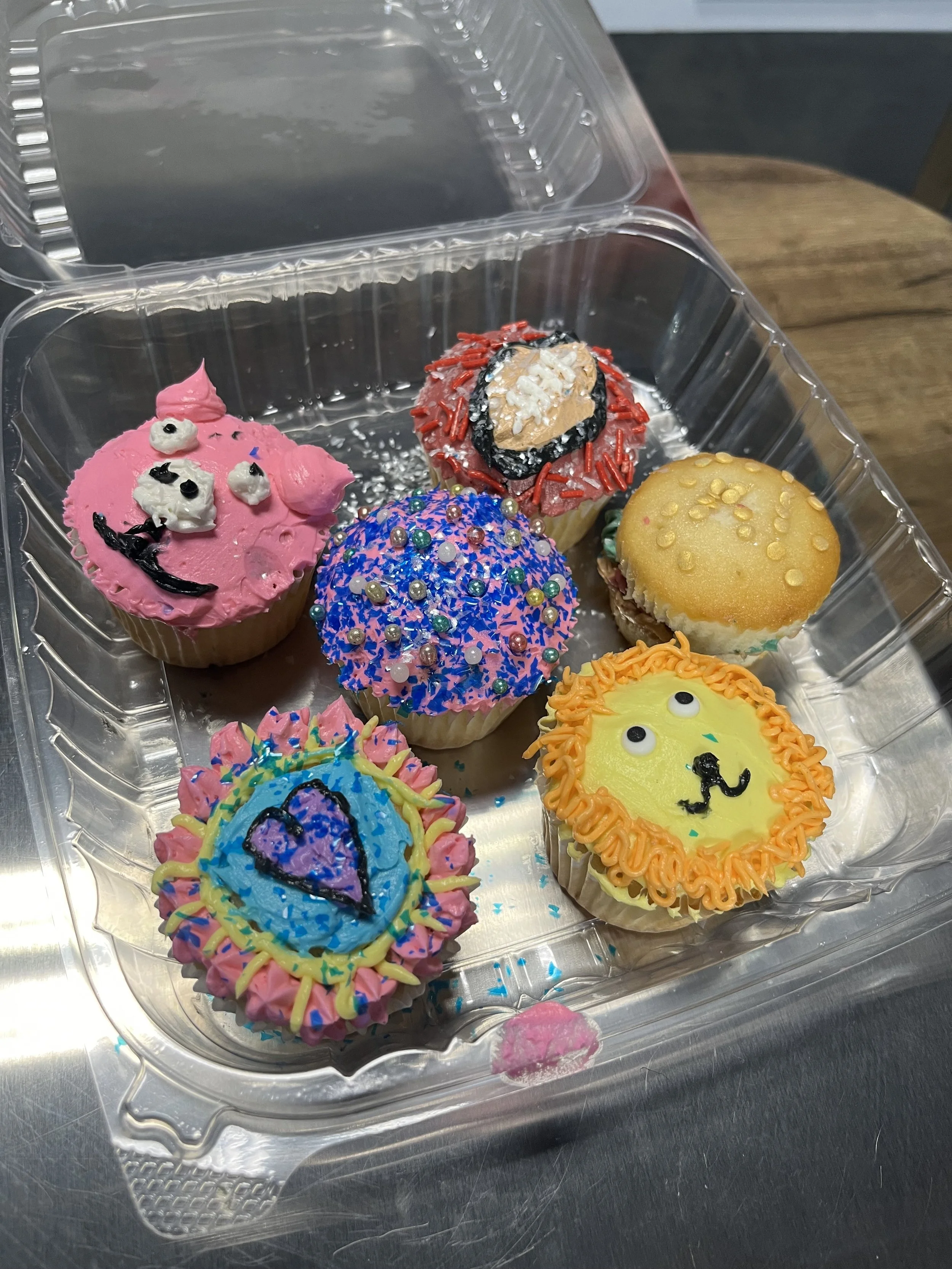 Set of decorated cupcakes with colorful frosting and sprinkles in a clear plastic container.