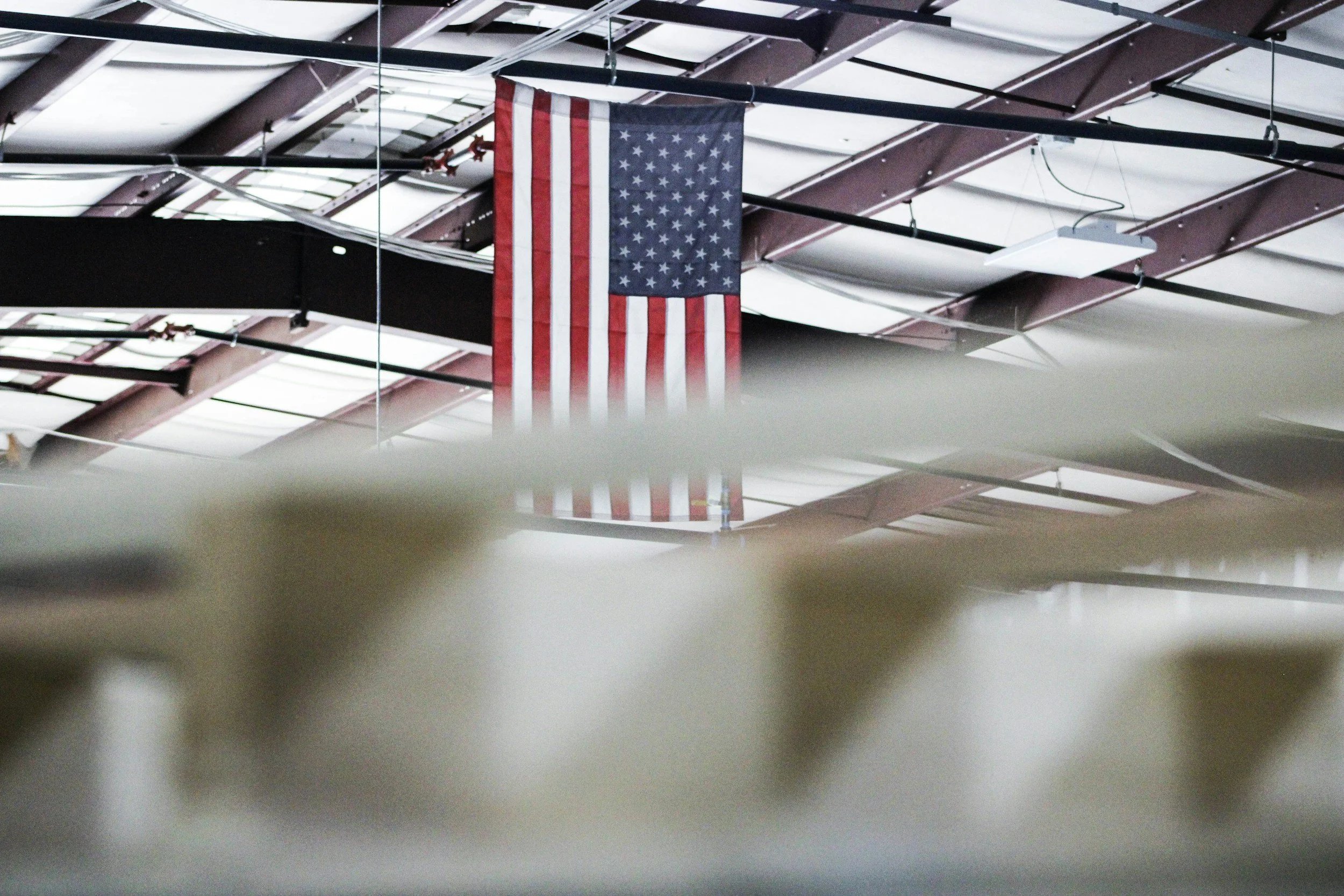 American flag hanging from the ceiling of an industrial or gymnasium space with a metal roof structure.