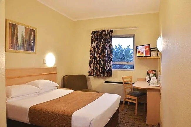 Hotel room with a bed, a window, a desk with a chair, a small shelf with picture frames, a couch, and wall art.