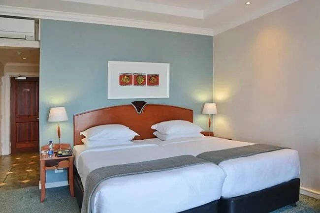 A hotel room with two beds pushed together, each with white bedding and gray runners. There are two nightstands with lamps on each side of the beds, and a picture with three pieces of art above the bed. The room has a blue accent wall and beige walls, with a door and an air conditioning unit visible.