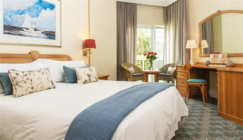 A cozy hotel room with a large bed, decorative pillows, and a blue throw blanket. There is a framed painting of a sailboat above the bed. A window with curtains overlooks a balcony with outdoor seating and flowers. Additional furniture includes a vanity with a mirror, a chair, and a small table with two chairs.