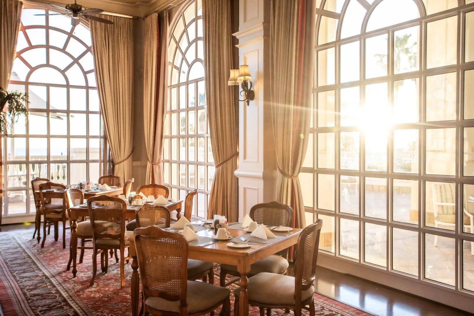 Elegant restaurant dining area with large arched windows letting in sunlight, wooden tables set with white napkins and china, surrounded by fruitwood chairs, and decorated with drapes and wall sconces.