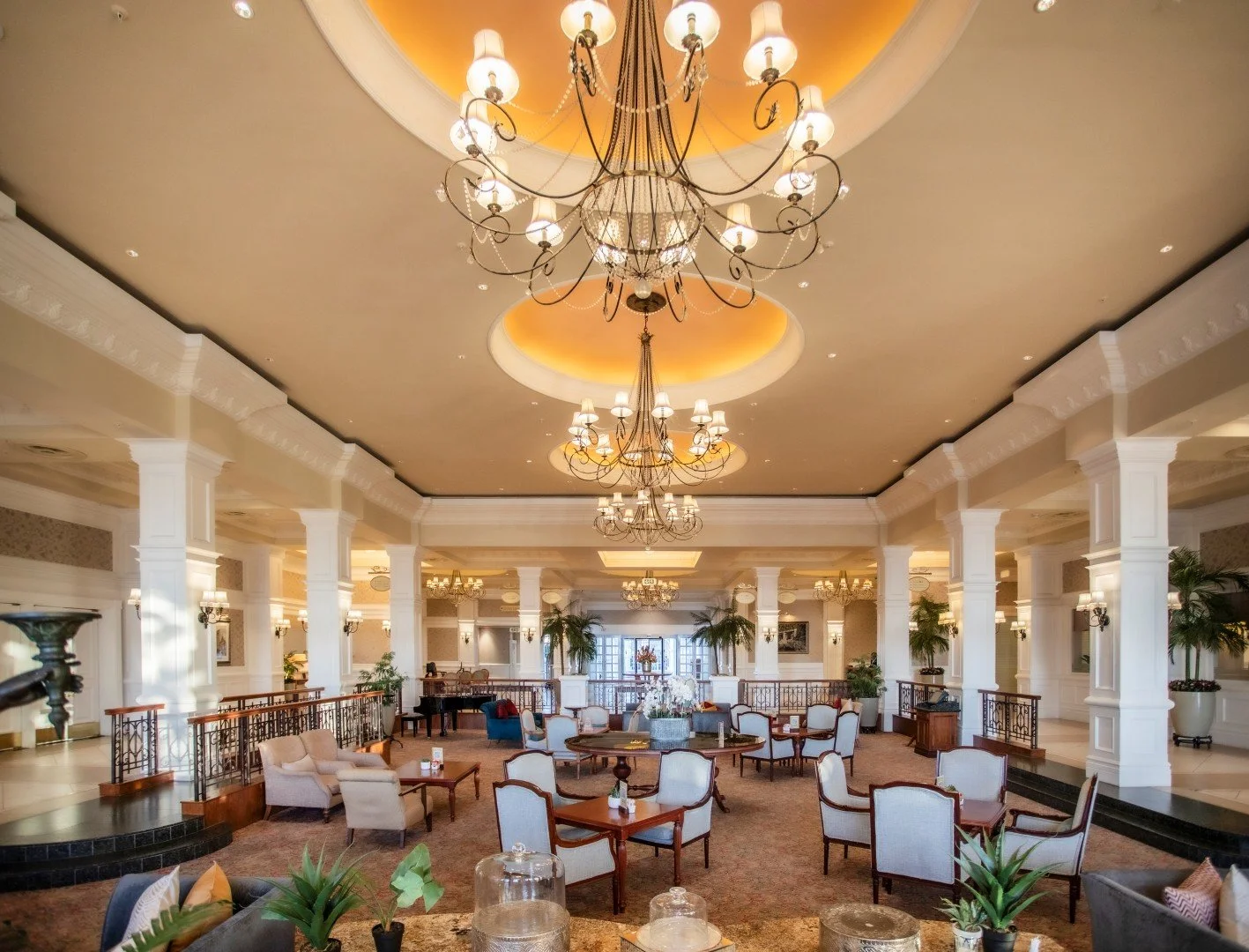 Elegant hotel lobby with high ceilings, large chandeliers, white columns, and seating areas with armchairs and small tables, decorated with plants and flowers.